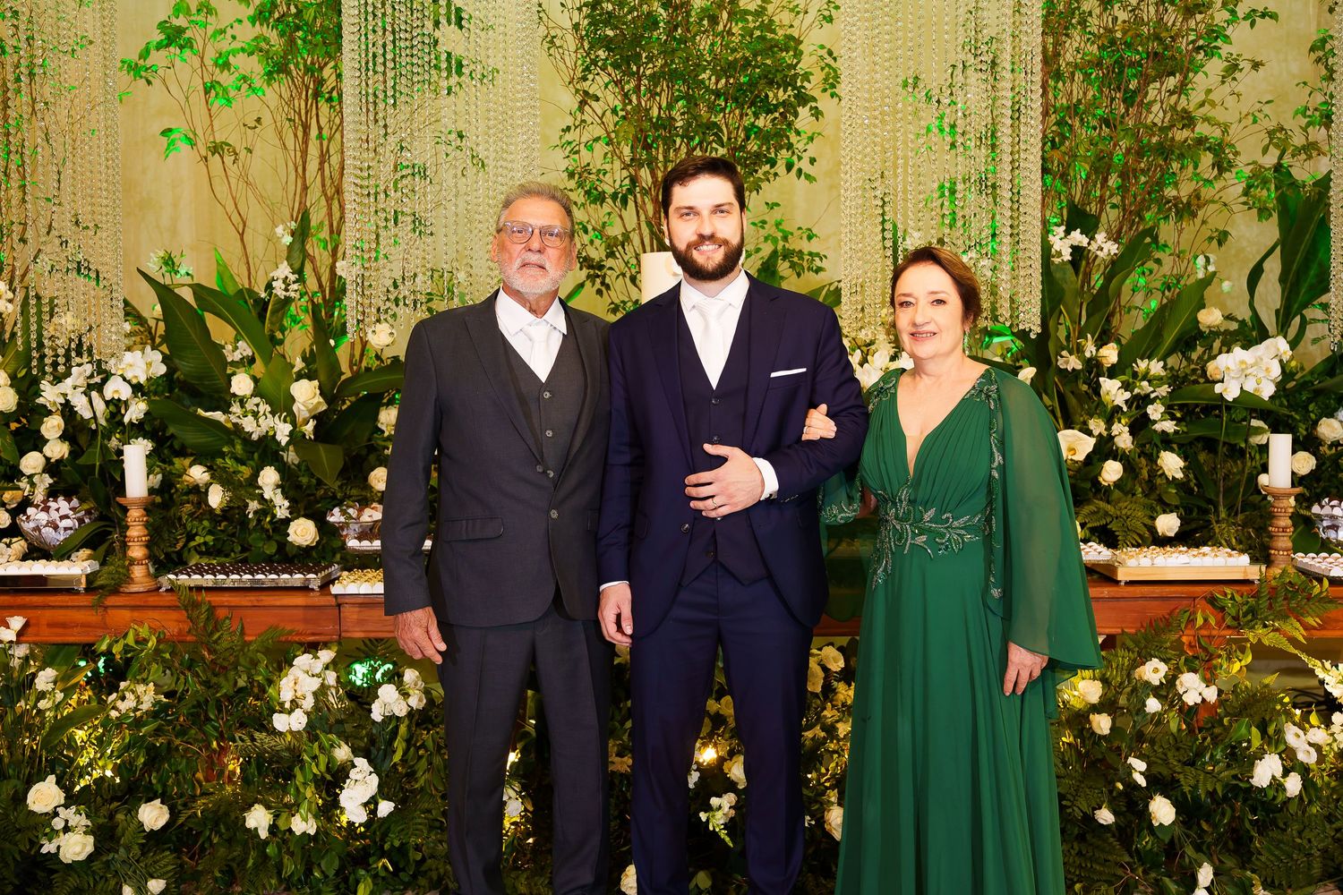 Three elegantly dressed people pose together at a formal event with lush green floral decorations.
