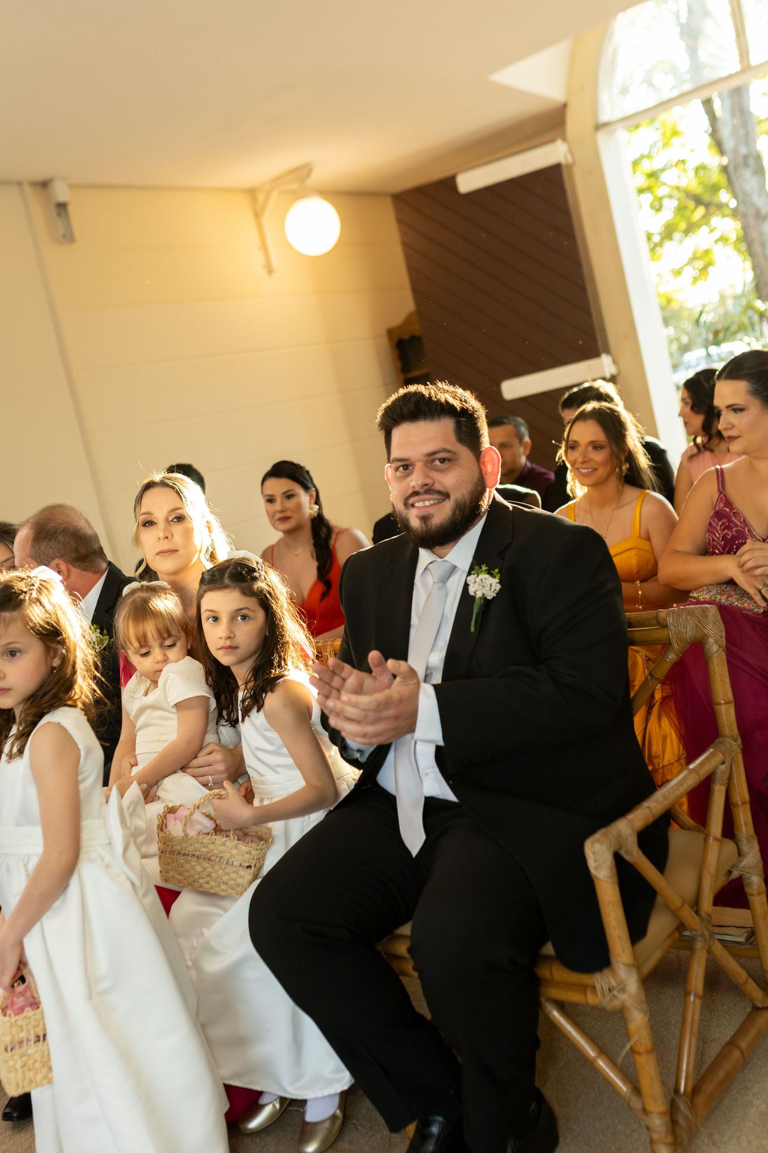 Groom in black tuxedo clapping at wedding ceremony surrounded by guests and flower girls in white dresses.