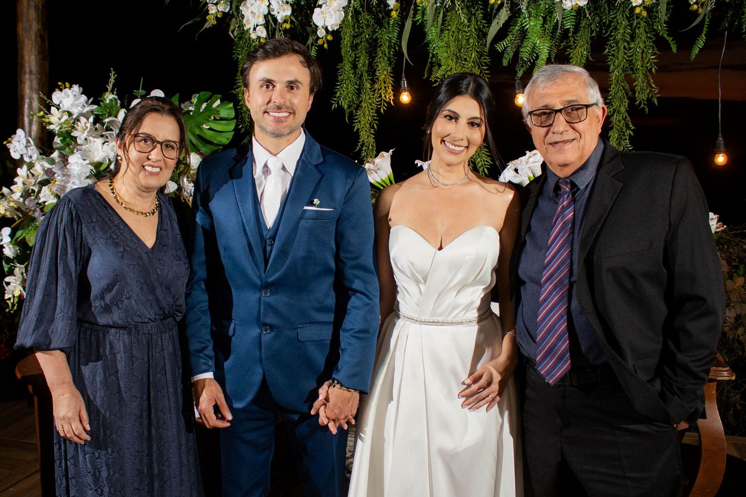 Bride and groom pose with two family members at an elegant wedding reception with floral arch backdrop.