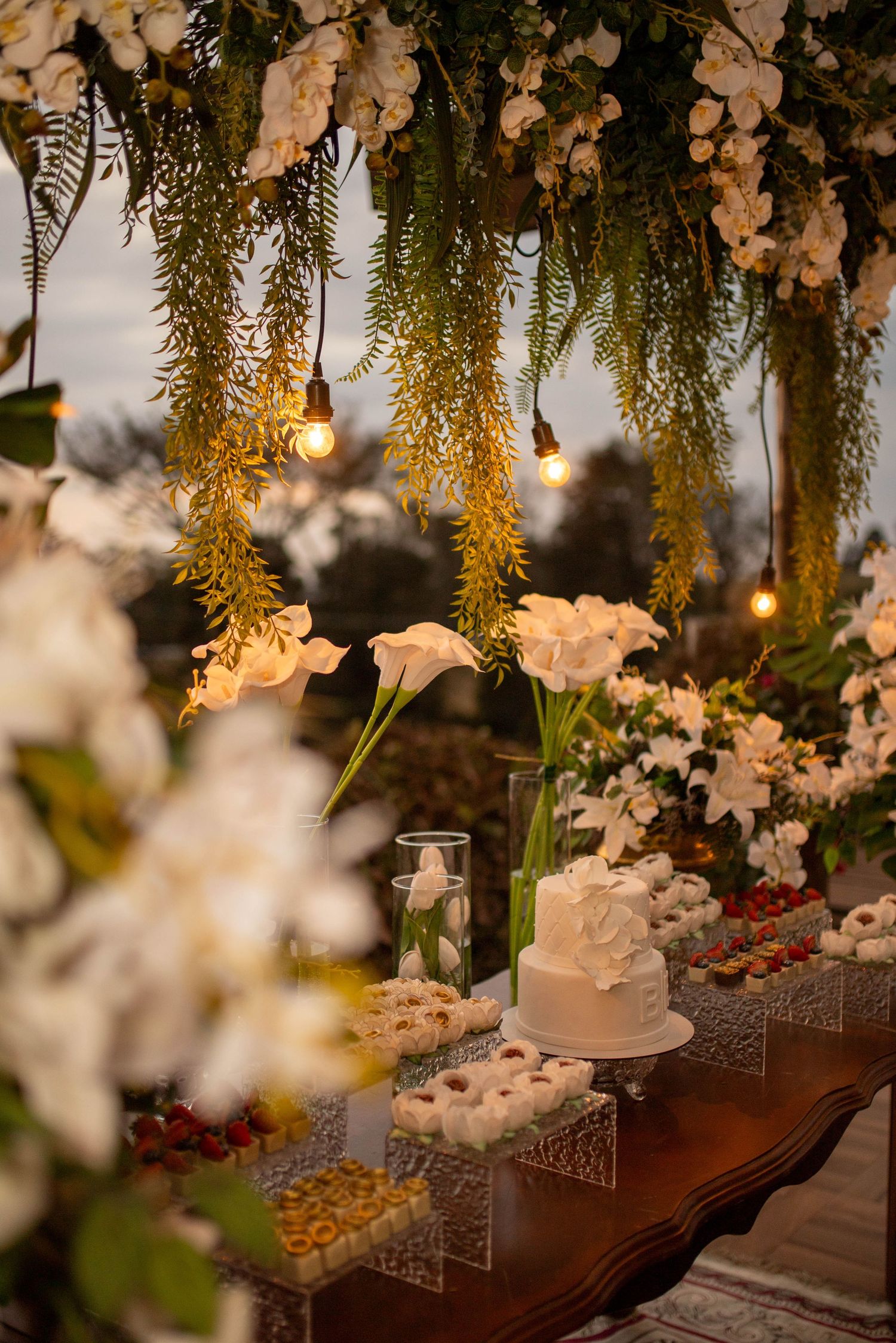 Elegant wedding dessert table with white cake, flowers, and warm Edison bulb lighting under floral canopy.