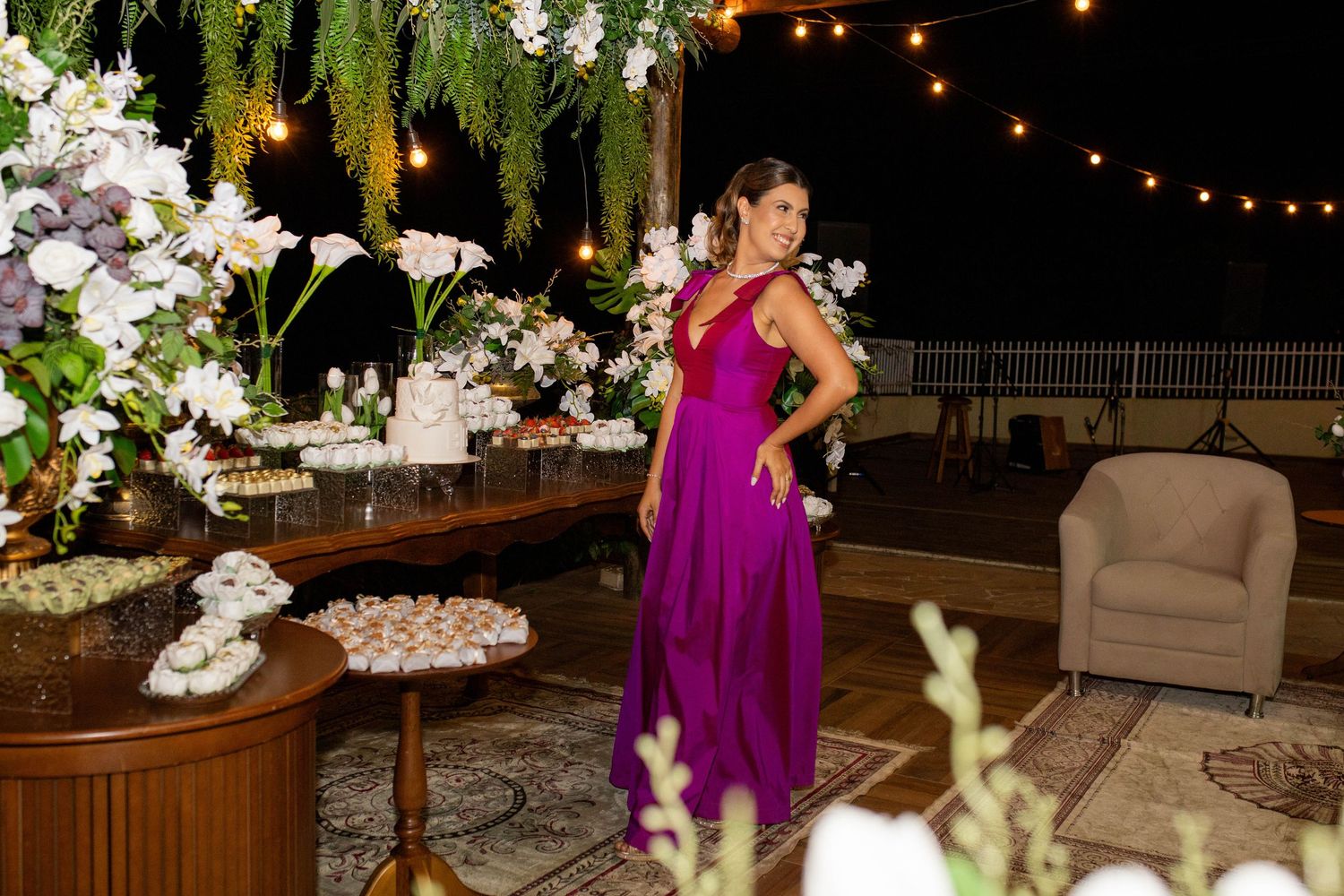 Woman in purple gown poses at elegant outdoor evening wedding reception with floral arrangements and string lights.