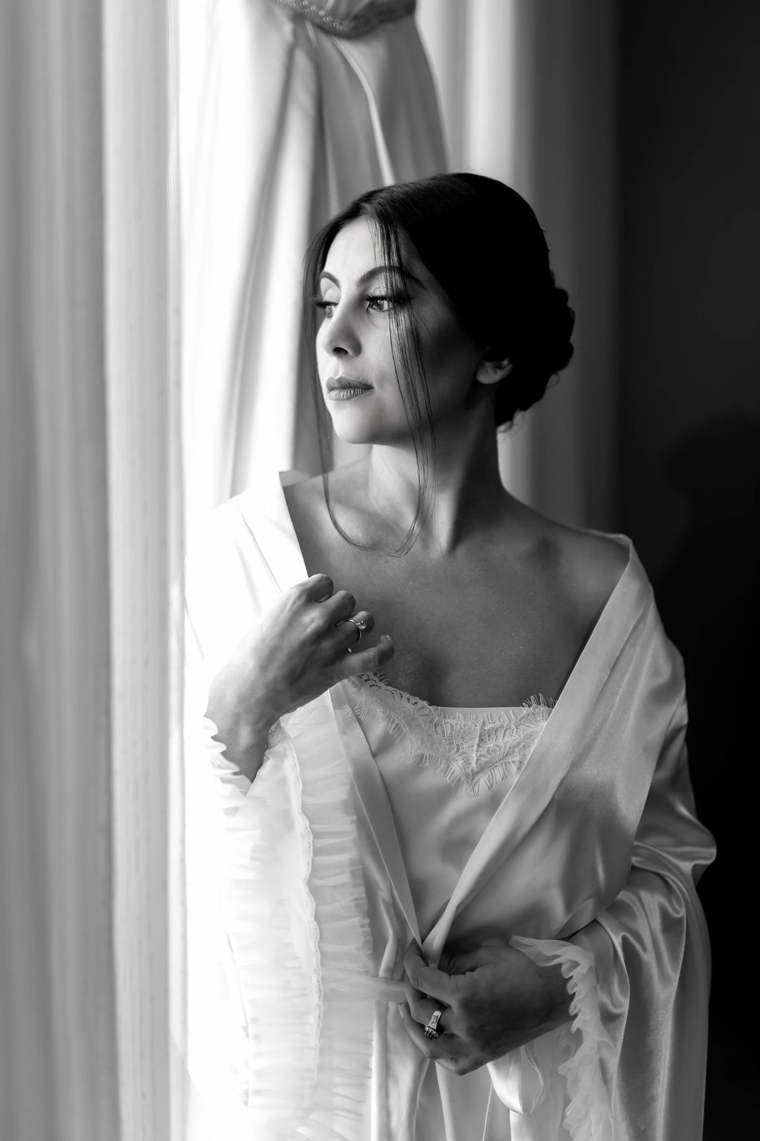 Black and white photo of an elegant bride in a satin robe gazing out a window, with soft natural light.