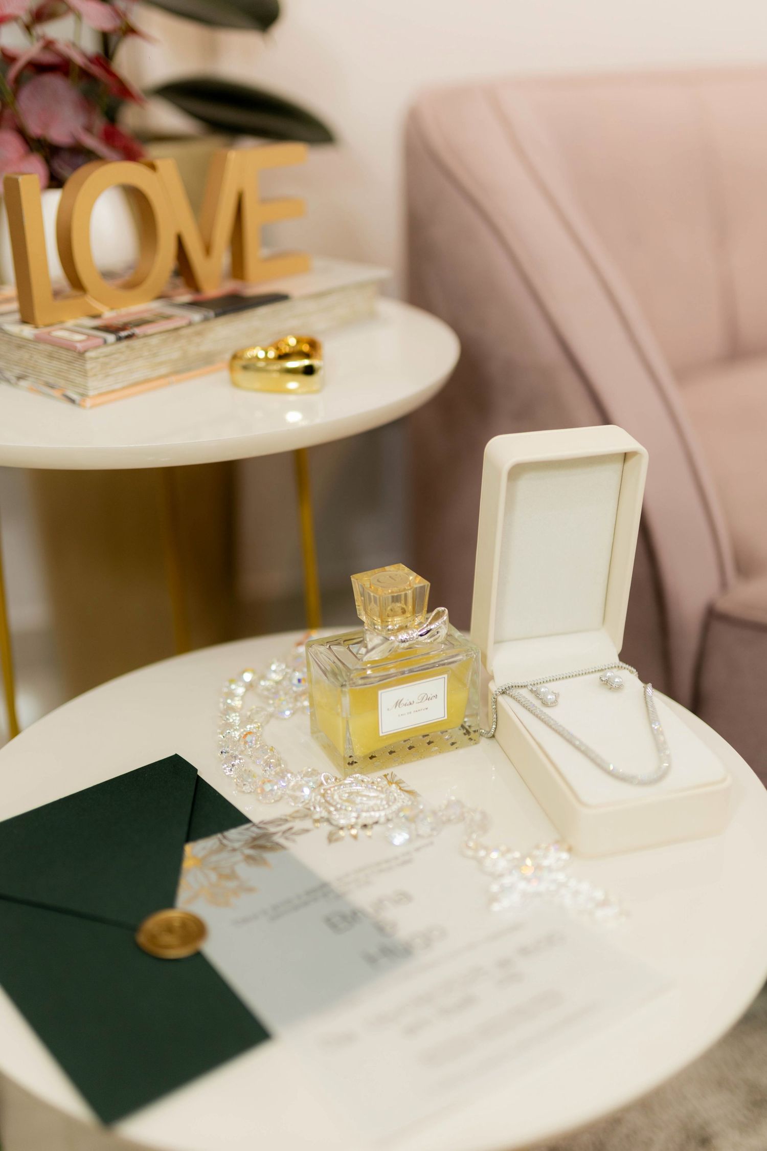 Wedding flat lay with Miss Dior perfume, pearl necklace, crystal hair piece, and green envelope.