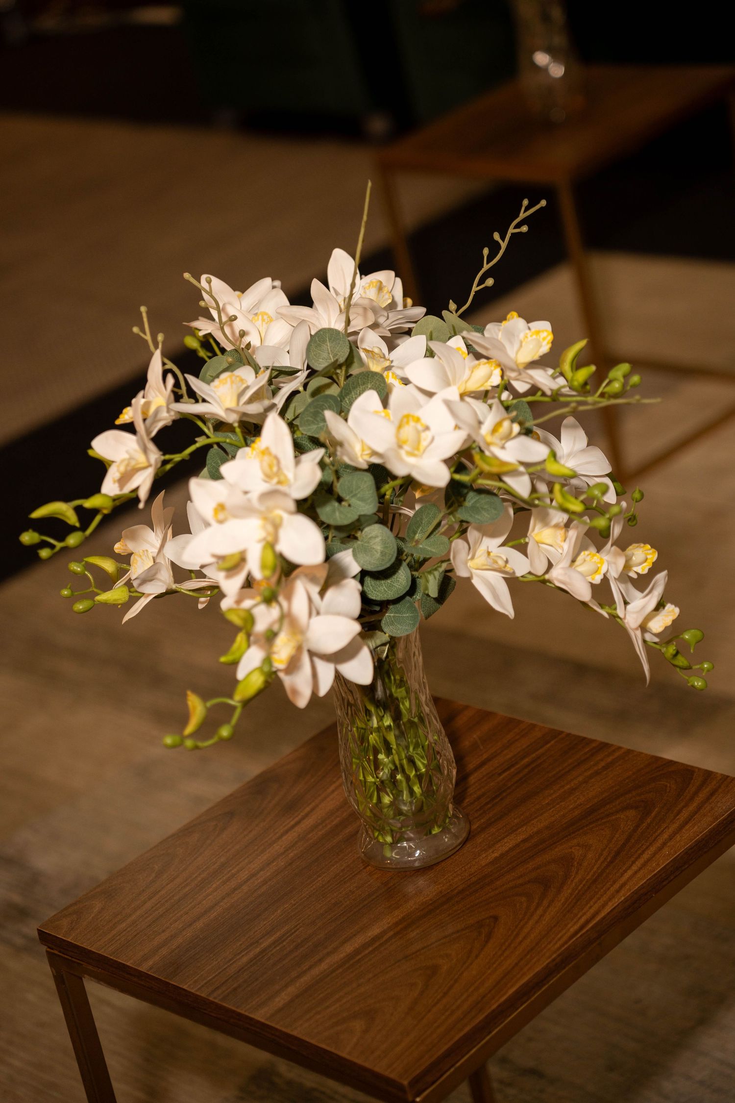 Elegant white orchid bouquet with green foliage in a glass vase on a wooden side table.