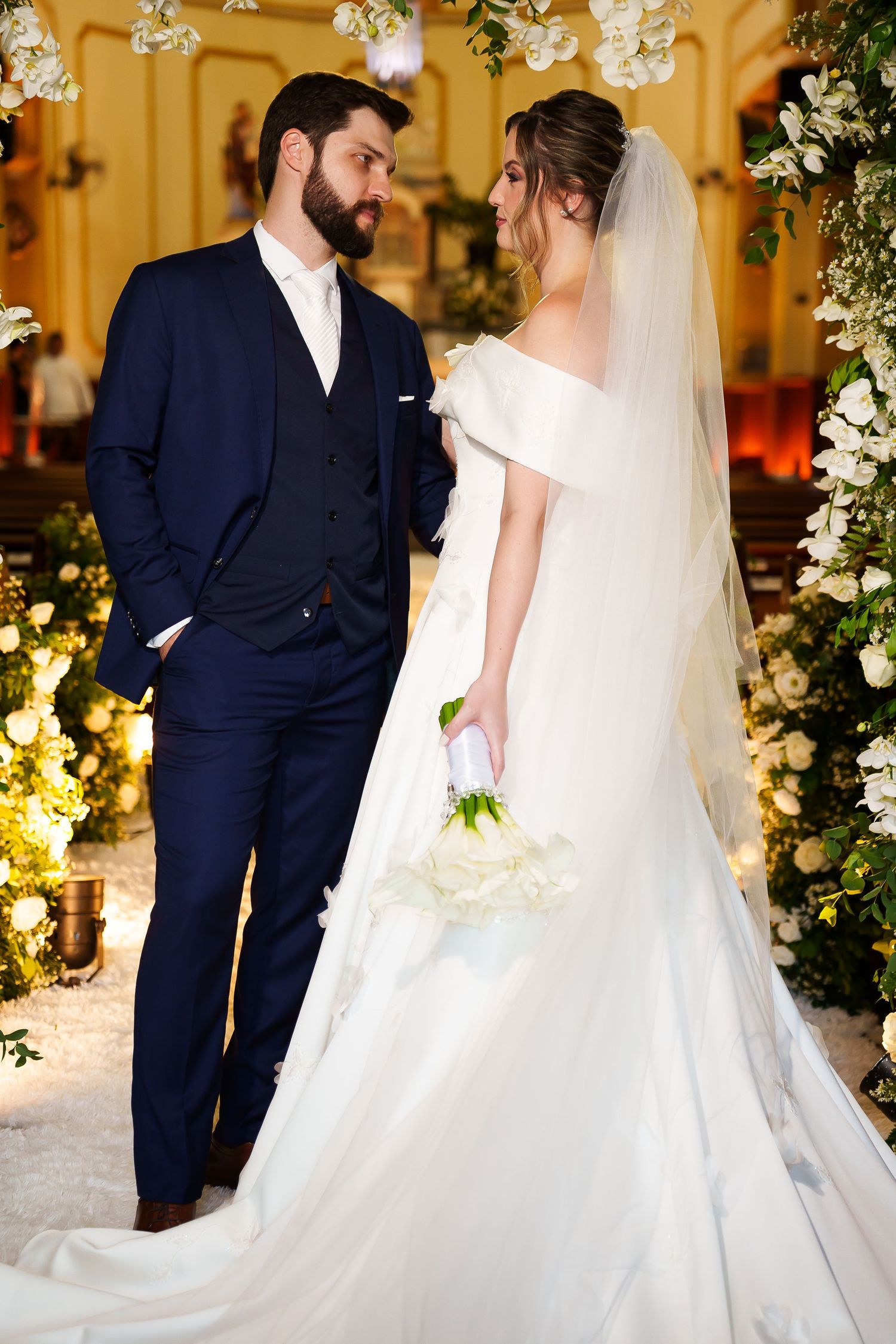 Bride in white off-shoulder gown holding bouquet stands with groom in navy suit inside decorated church.