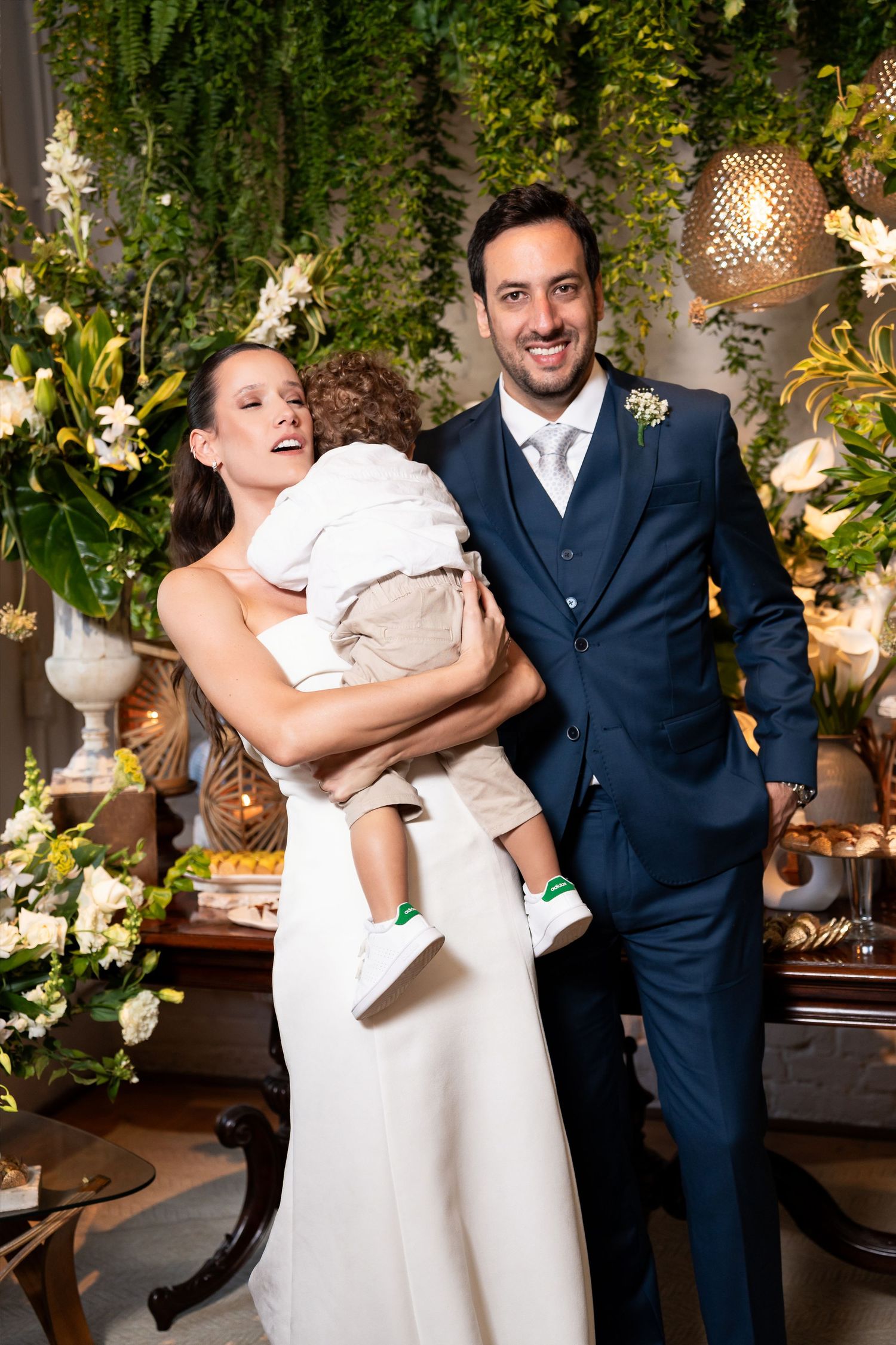 Bride in white gown holds young child beside groom in navy suit at floral wedding reception.