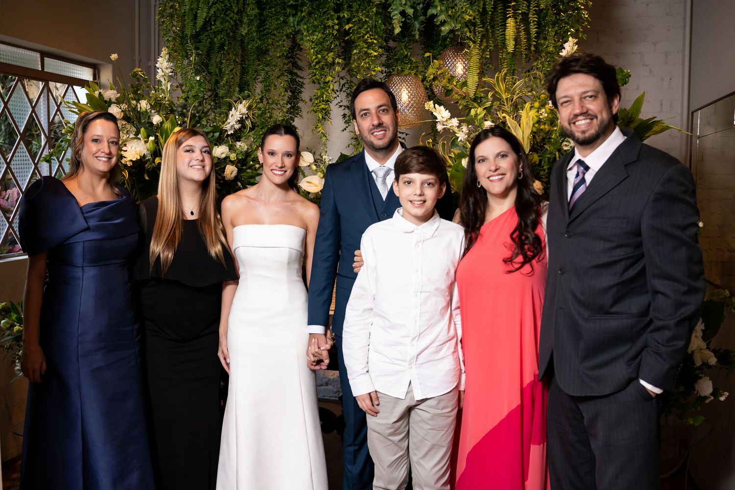 Wedding couple poses with family members in formal attire before a lush green floral backdrop.