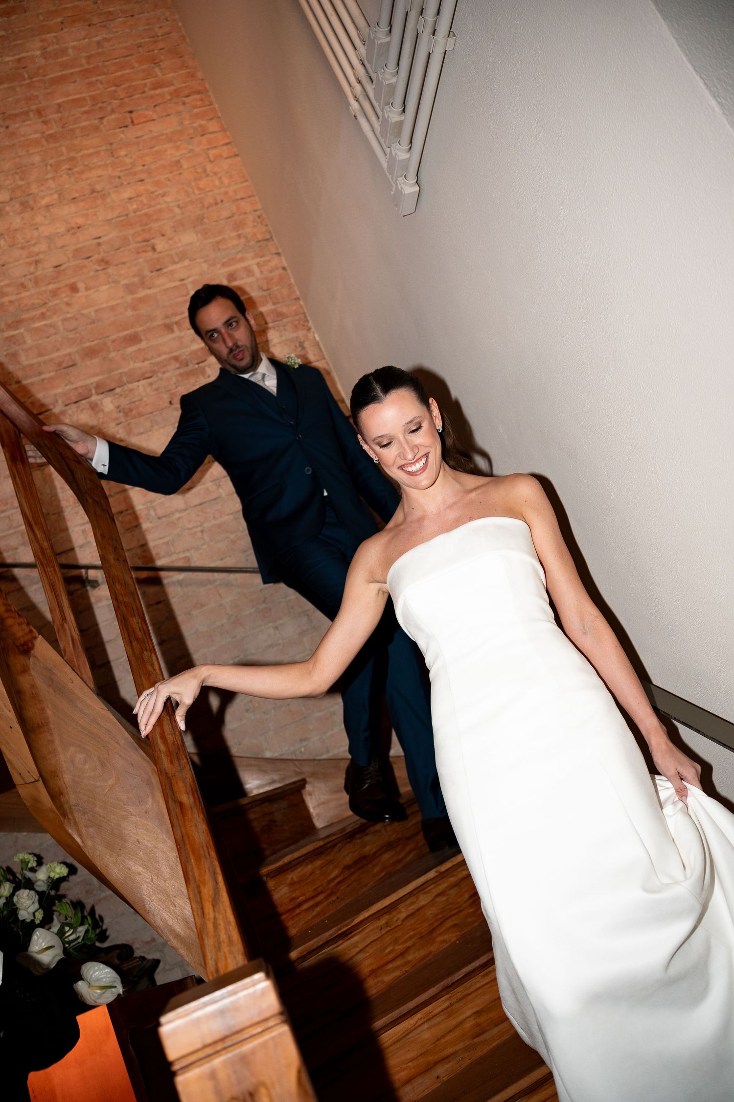 Bride in white strapless gown and groom in navy suit descending wooden staircase, smiling during wedding reception.