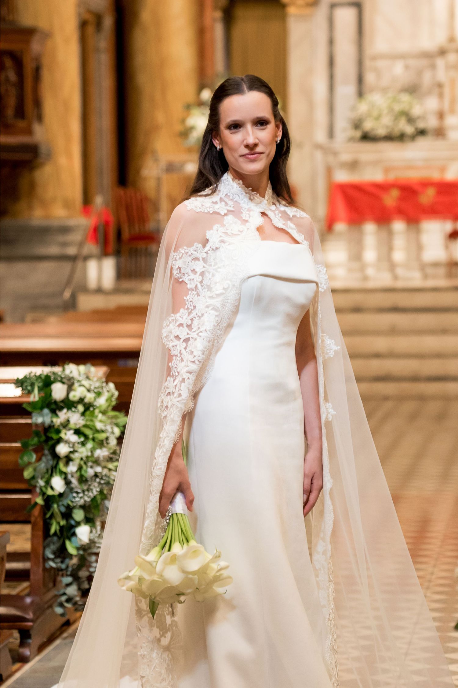 Bride in elegant white wedding gown with lace cape veil holding calla lily bouquet inside ornate church.
