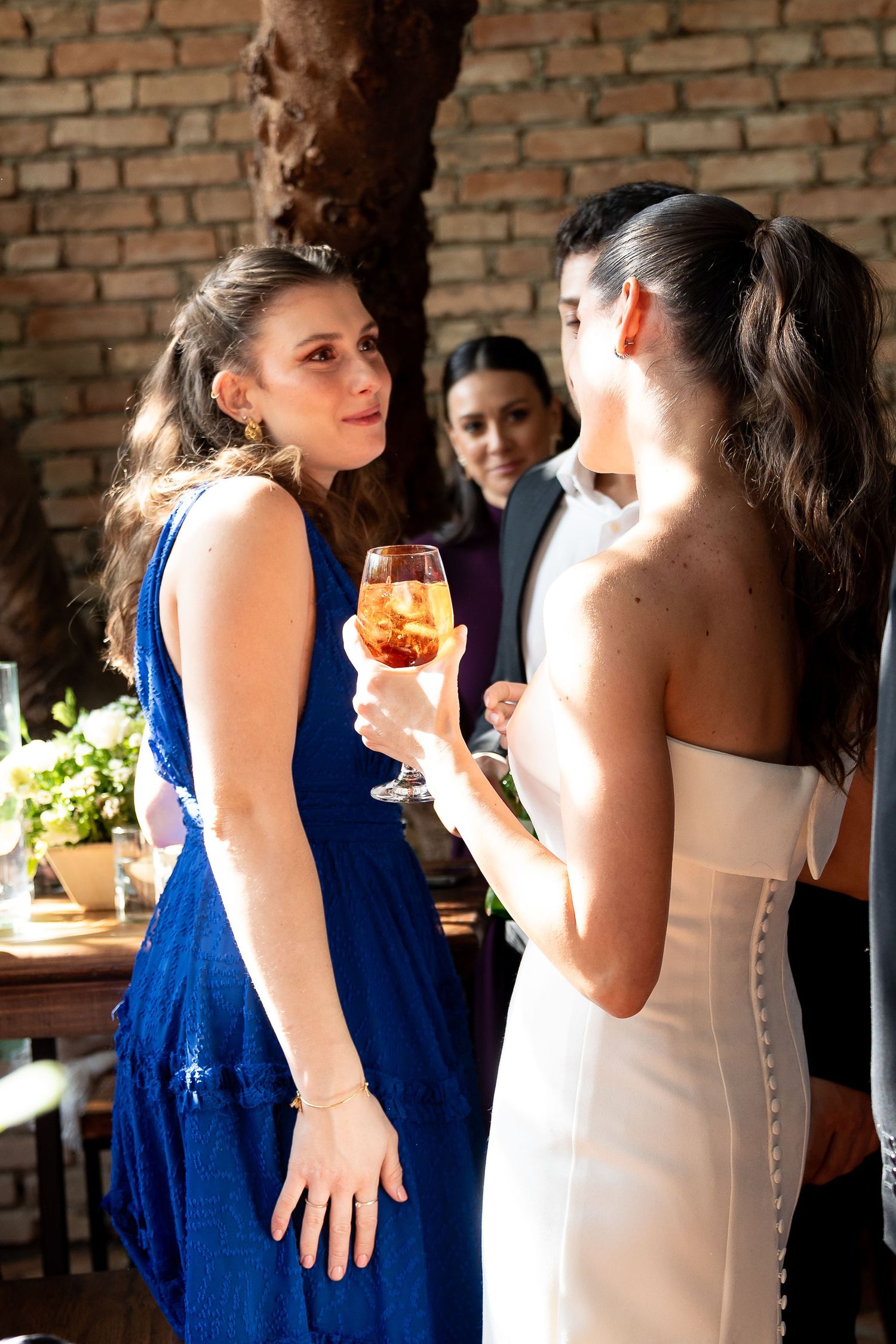 Two women in elegant dresses chat at a wedding reception, one in blue holding a cocktail glass.
