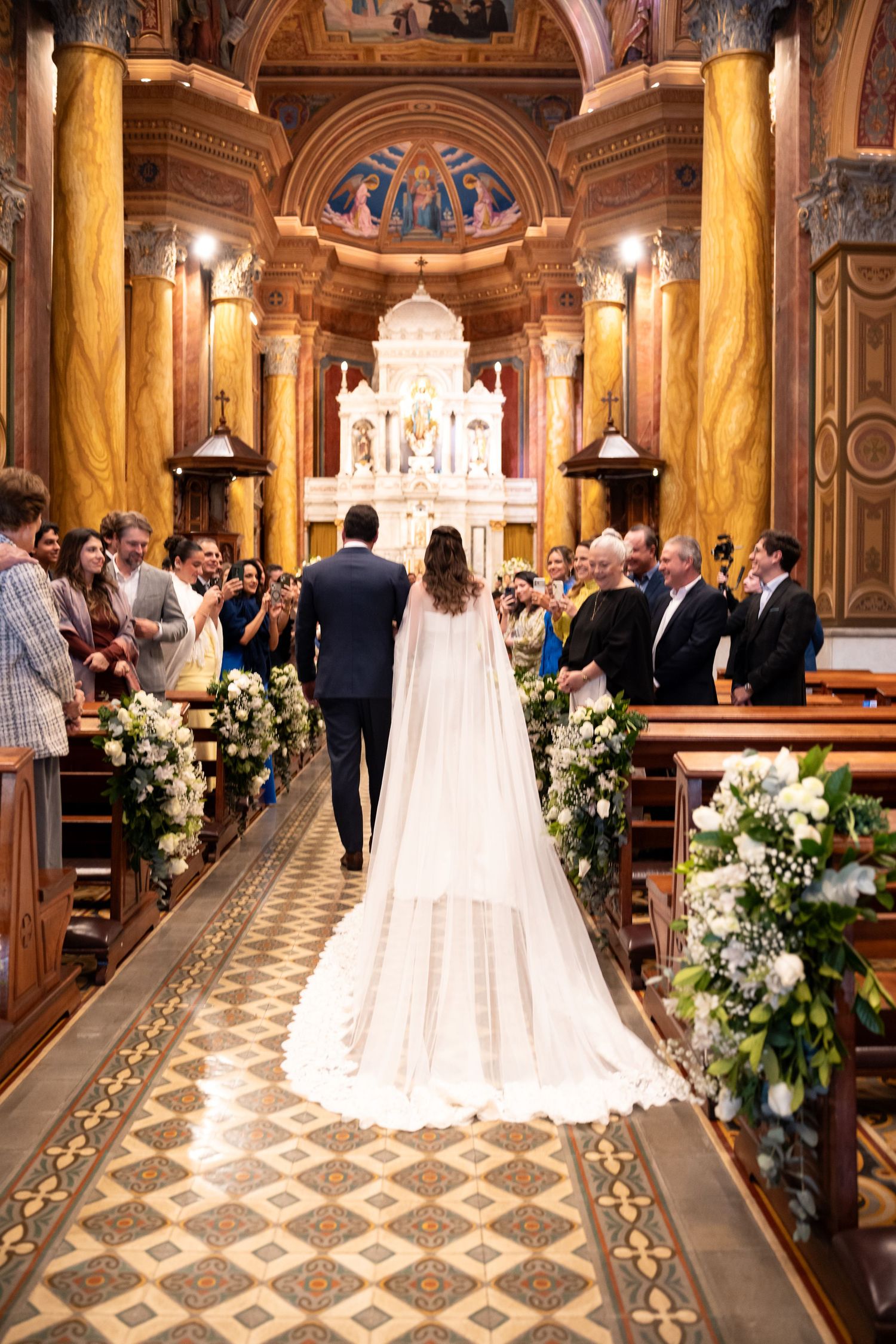 Bride and groom walking down the aisle of an ornate church with floral arrangements and guests during a wedding ceremony.
