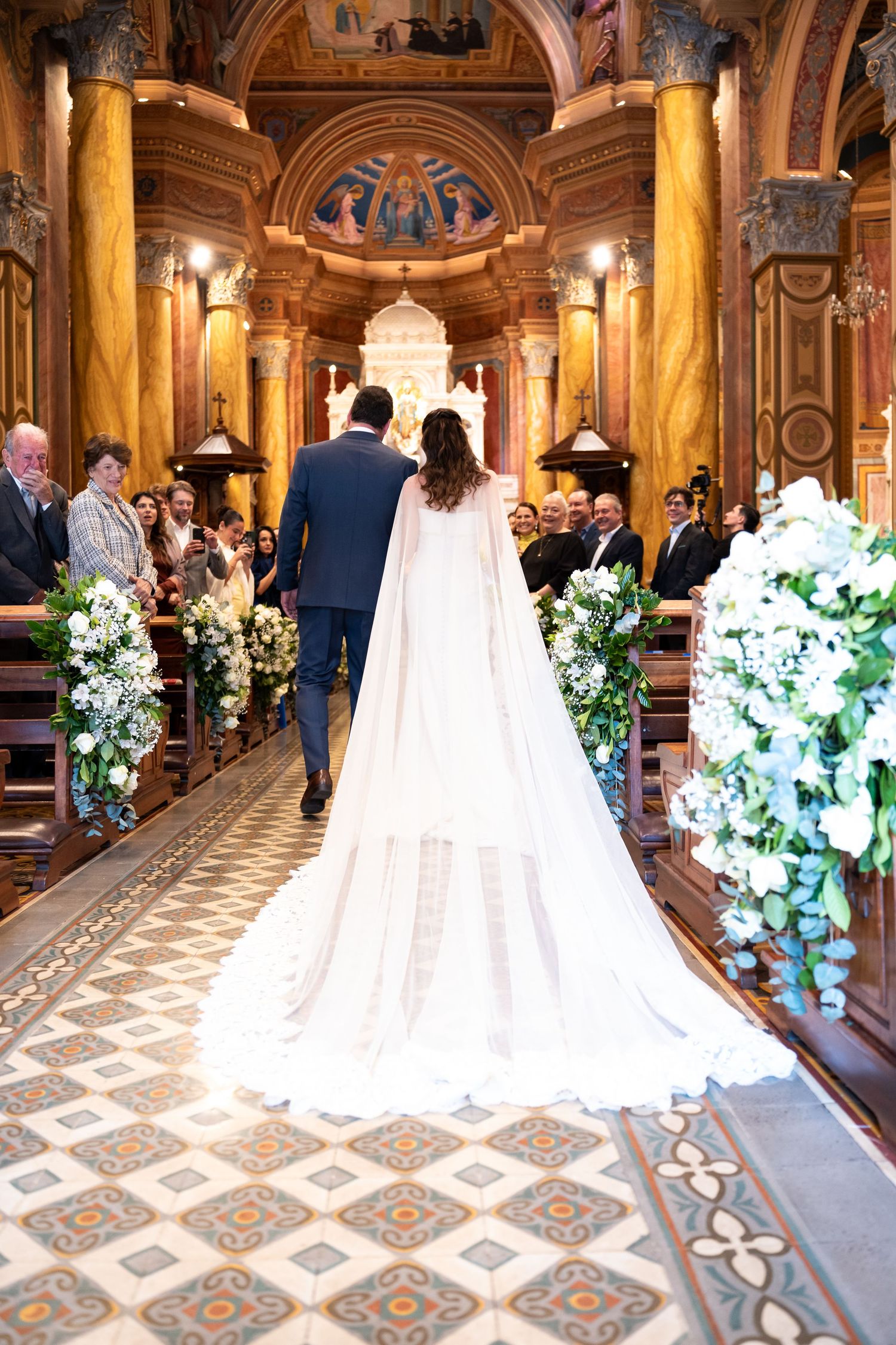 Bride and groom walking down the aisle of an ornate church decorated with white floral arrangements during wedding ceremony.