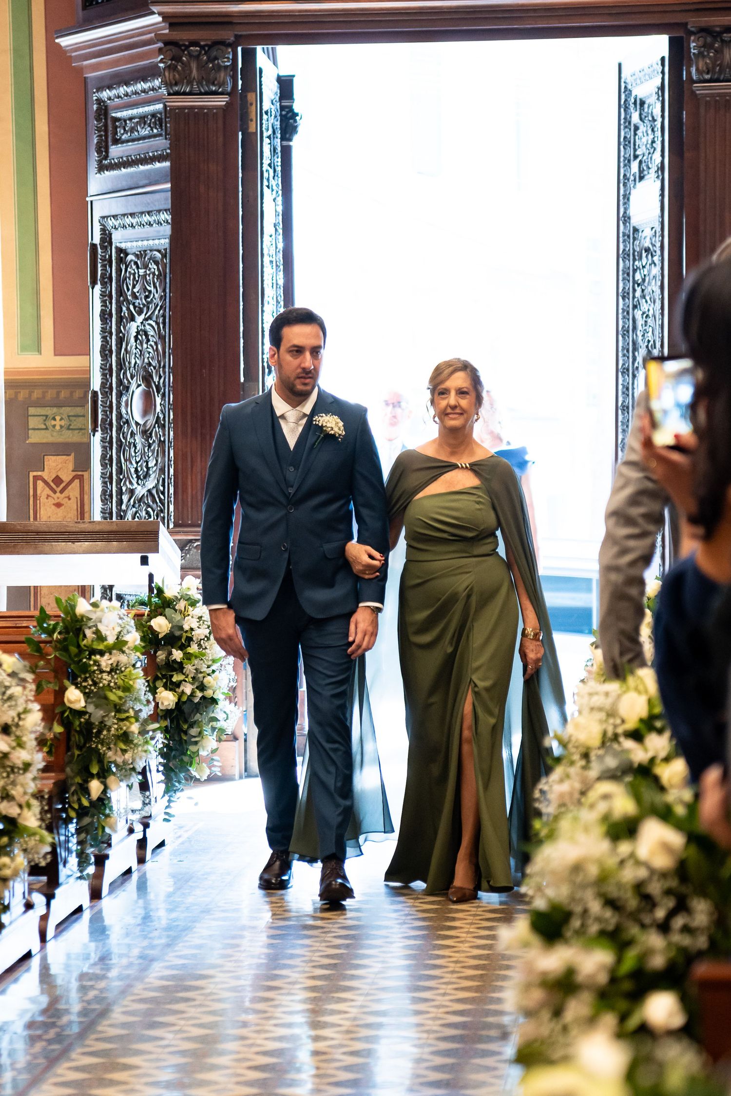 Groom in navy suit escorts woman in olive green gown down flower-lined church aisle at elegant wedding ceremony.