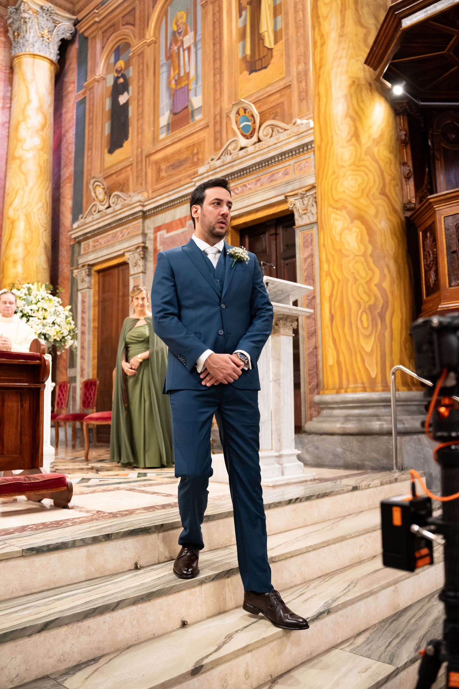 Groom in navy blue suit standing at church altar with ornate golden frescoes and marble columns.
