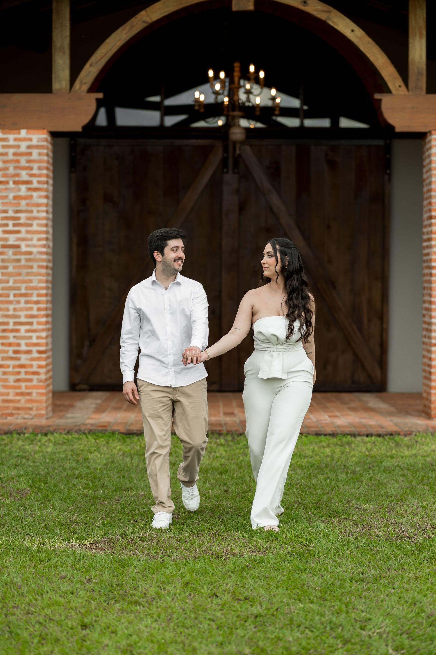 Couple holding hands walking on green grass in front of rustic barn doors with brick archway and chandelier.