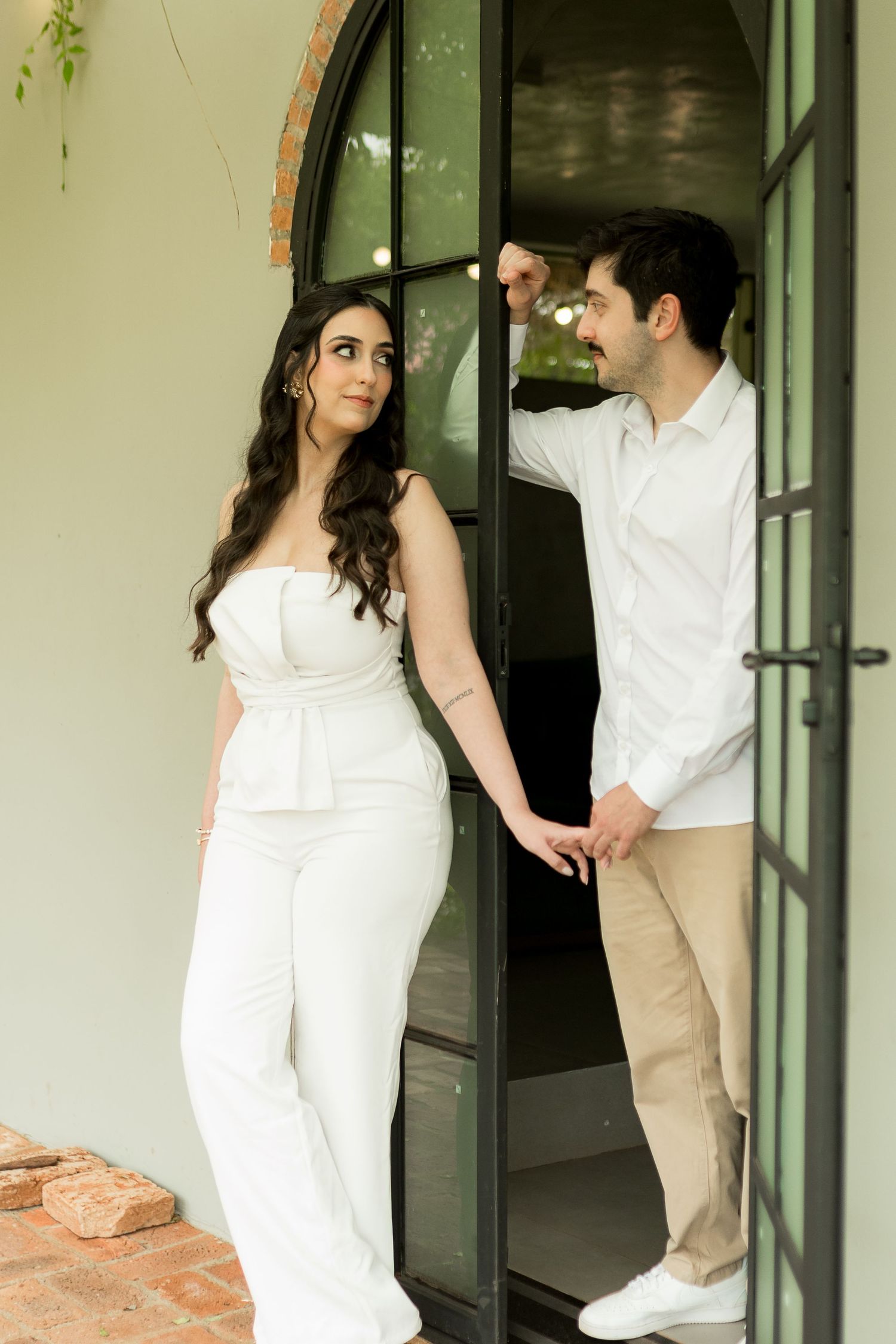 Couple in white outfits holding hands by an arched glass door, woman in white jumpsuit and man in white shirt.