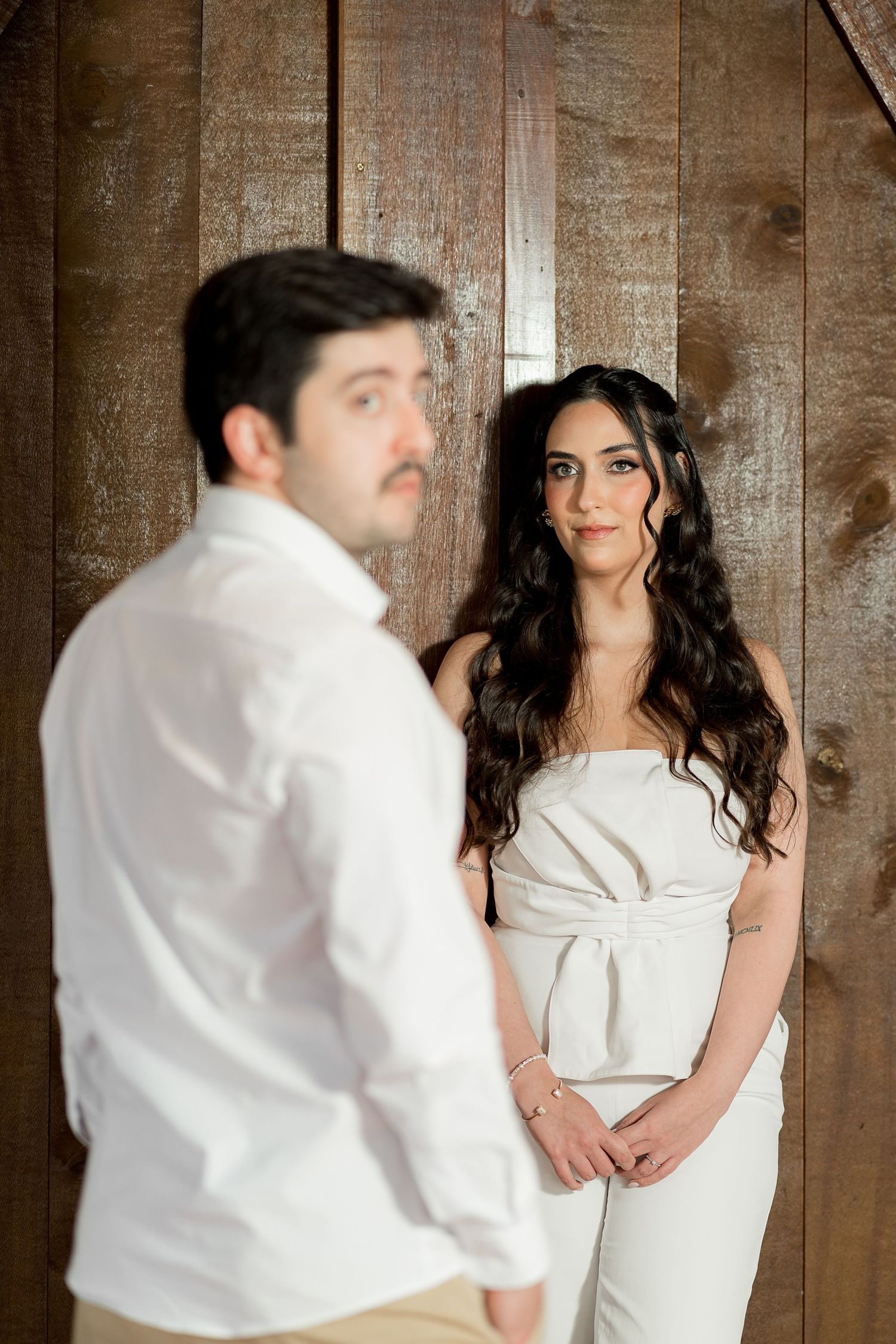 A bride in a white strapless gown gazes at a man in a white shirt against a rustic wooden wall backdrop.