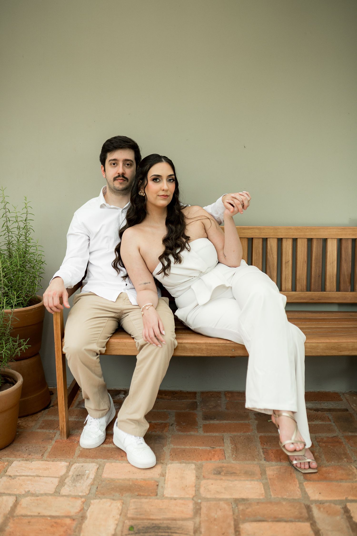 Couple in white outfits sitting together on a wooden bench against a sage green wall on a brick patio.