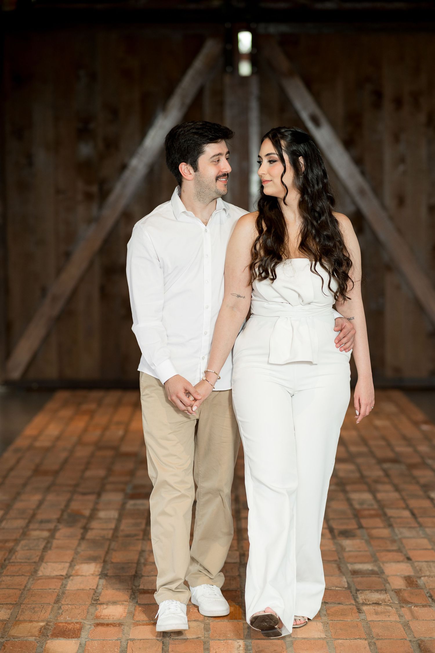 A couple in white and beige outfits hold hands and smile at each other inside a rustic barn.