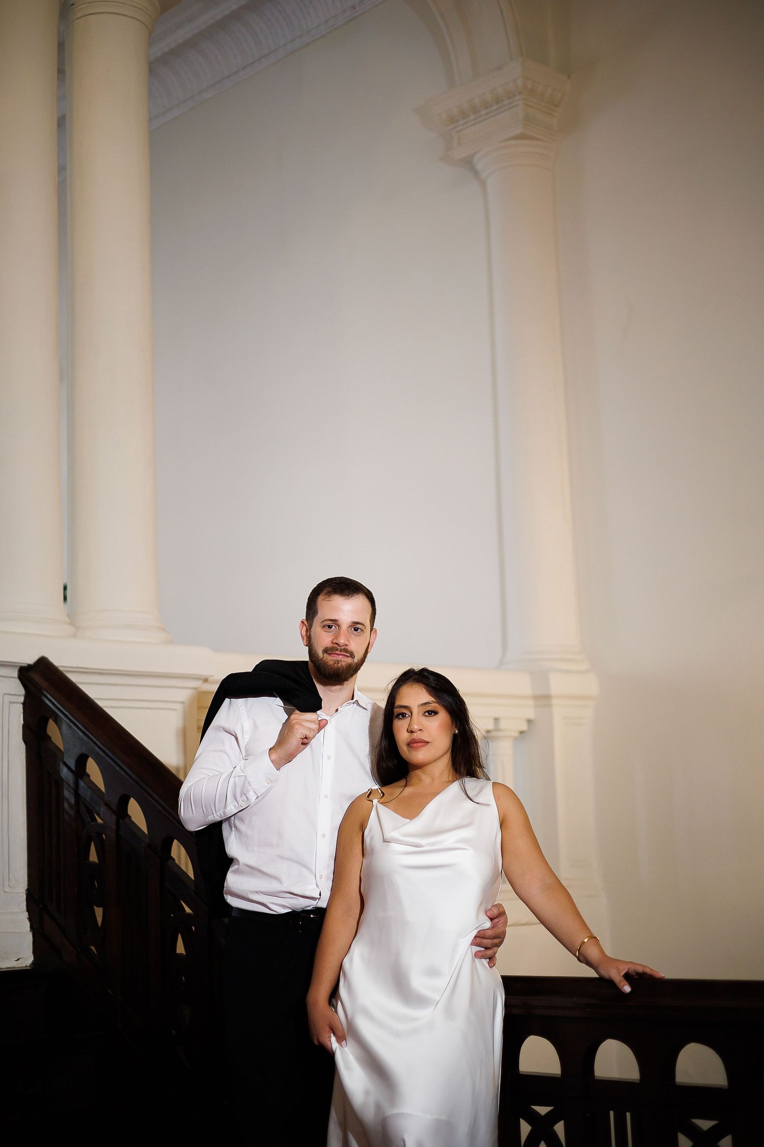Elegant couple posing on ornate wooden staircase, woman in white satin dress, man in white shirt holding black jacket.