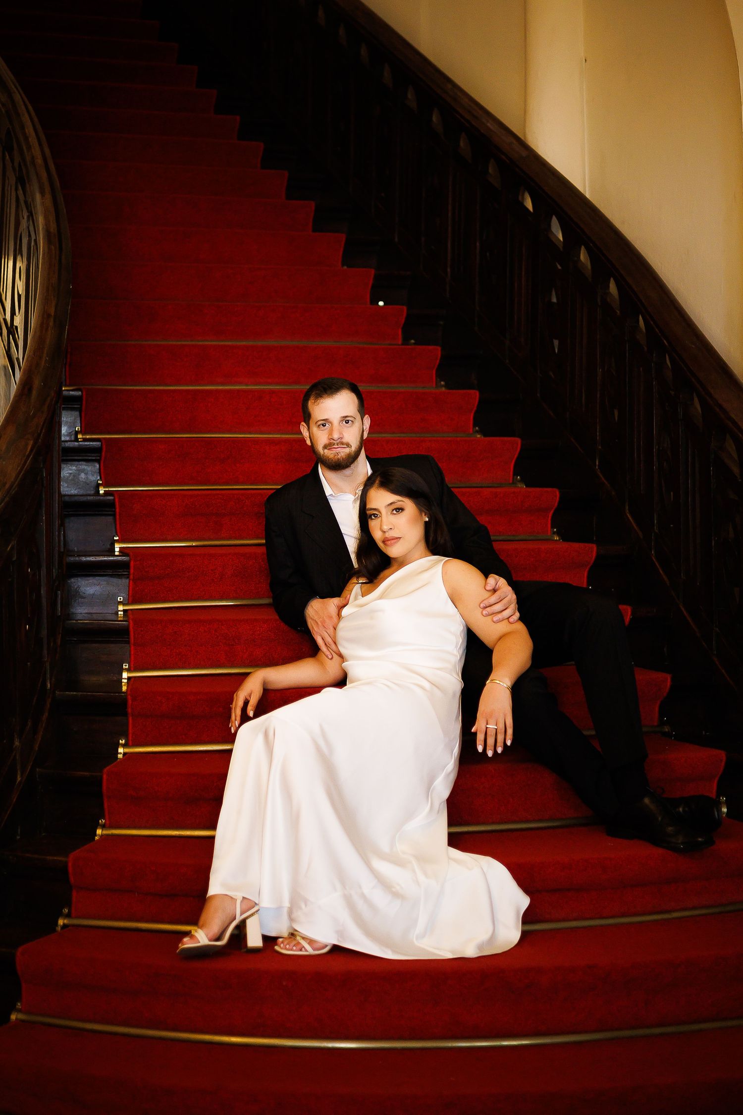 Bride in white gown and groom in black suit pose elegantly on red carpeted staircase.