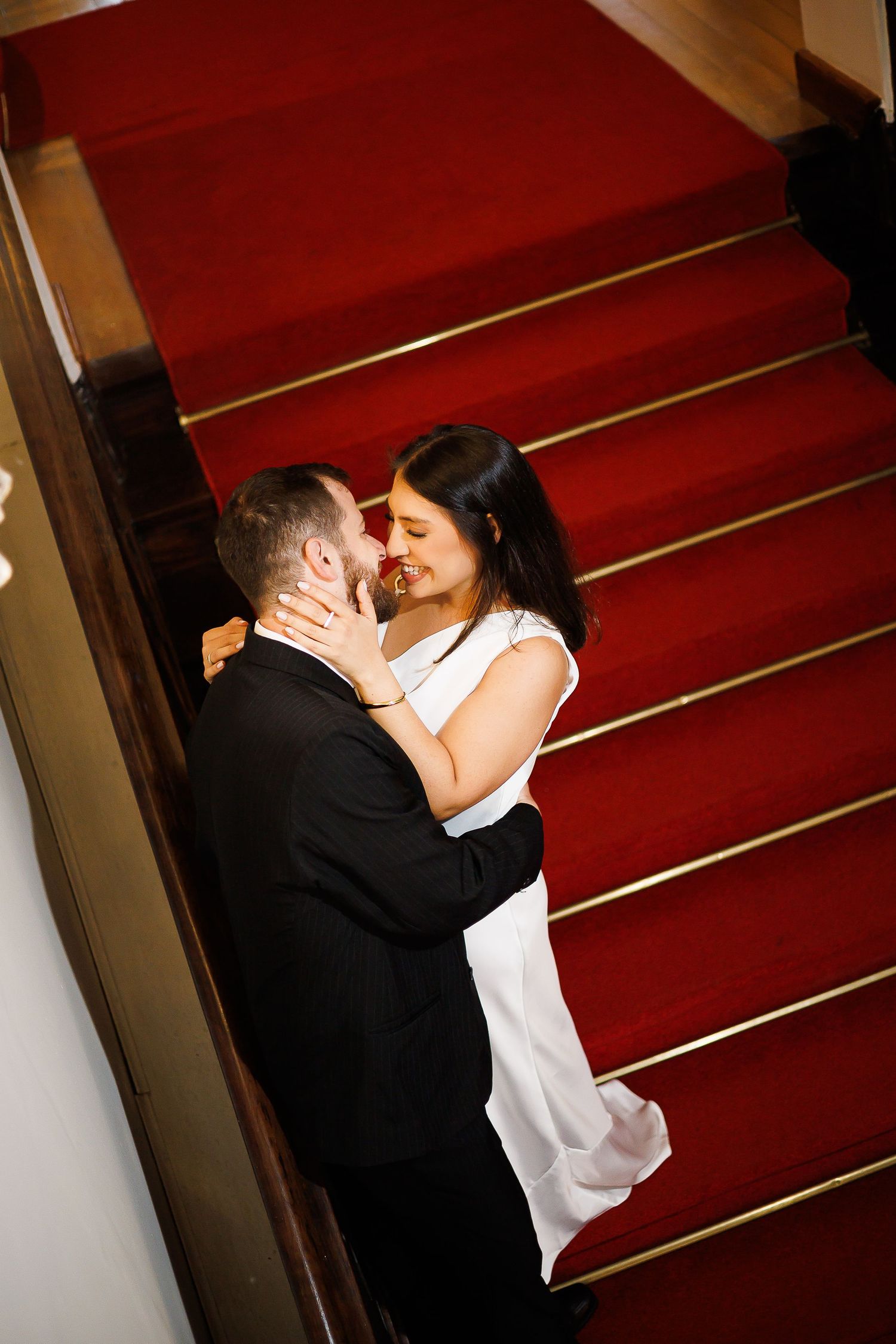 Couple sharing a romantic moment at the base of a grand staircase with red carpet, bride in white dress.
