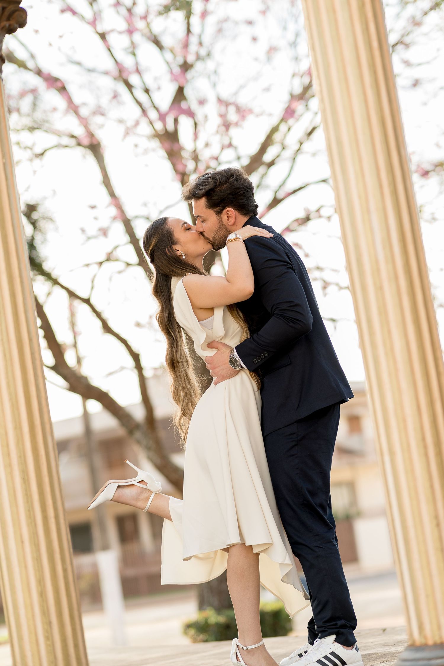 Couple sharing a romantic kiss between white columns with pink cherry blossom trees in background during engagement photo.