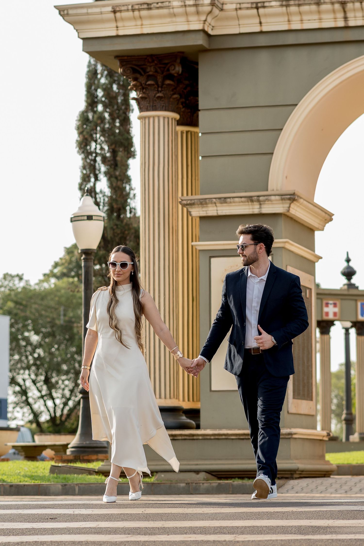 Elegant couple holding hands walking past a classical arch monument, woman in white dress and man in navy suit.