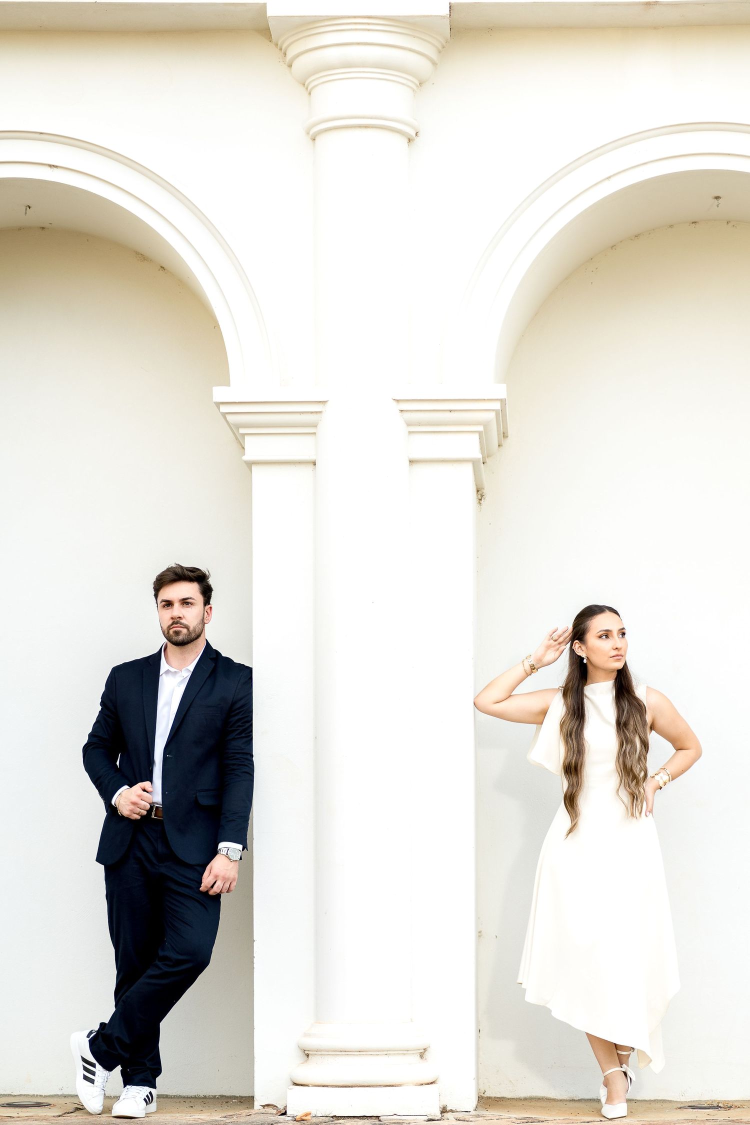 Elegant couple posing by white arched columns, man in dark suit, woman in white dress with long hair.