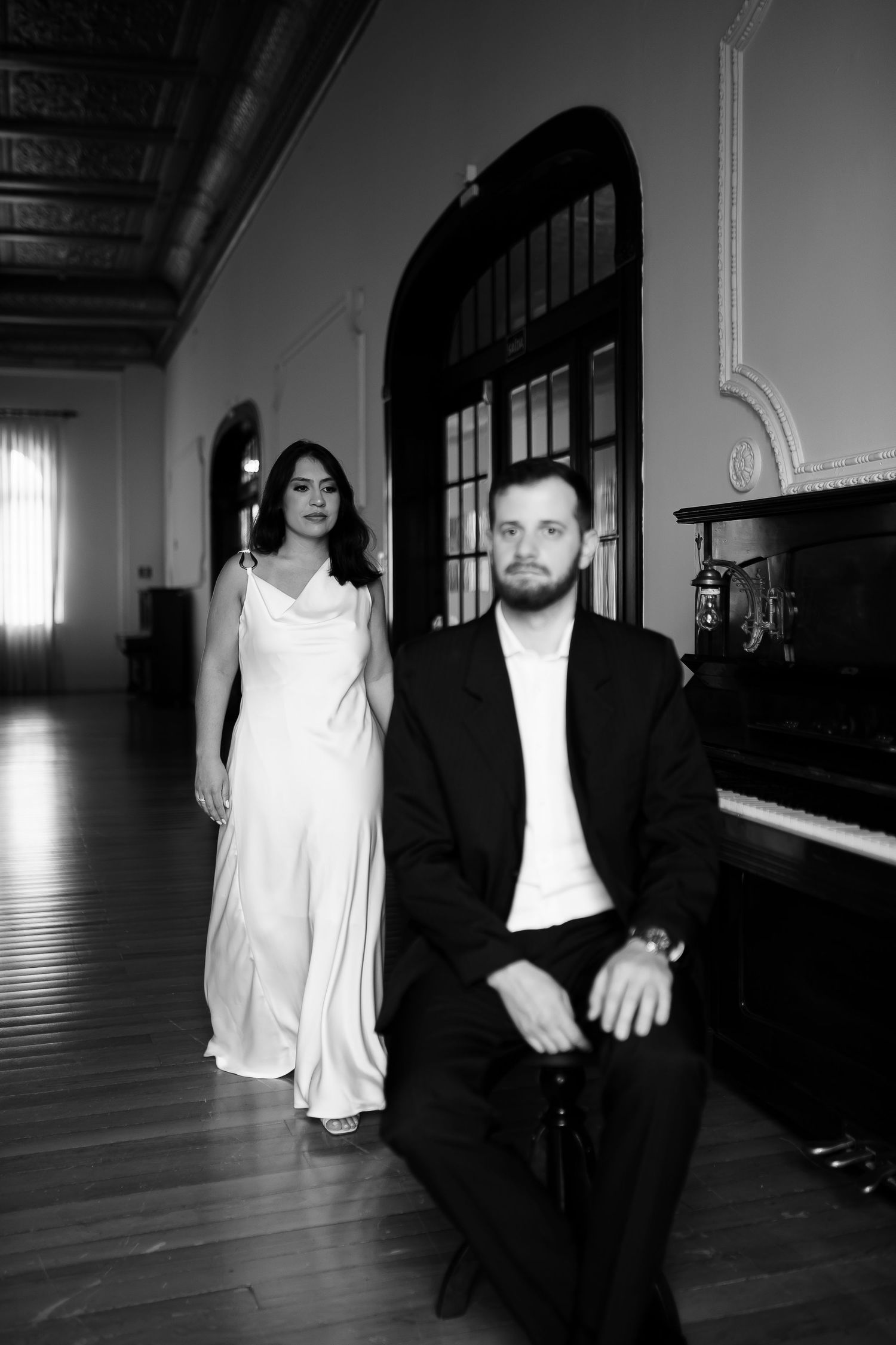 Black and white photo of a woman in white dress and bearded man in suit seated near a grand piano in elegant hall.