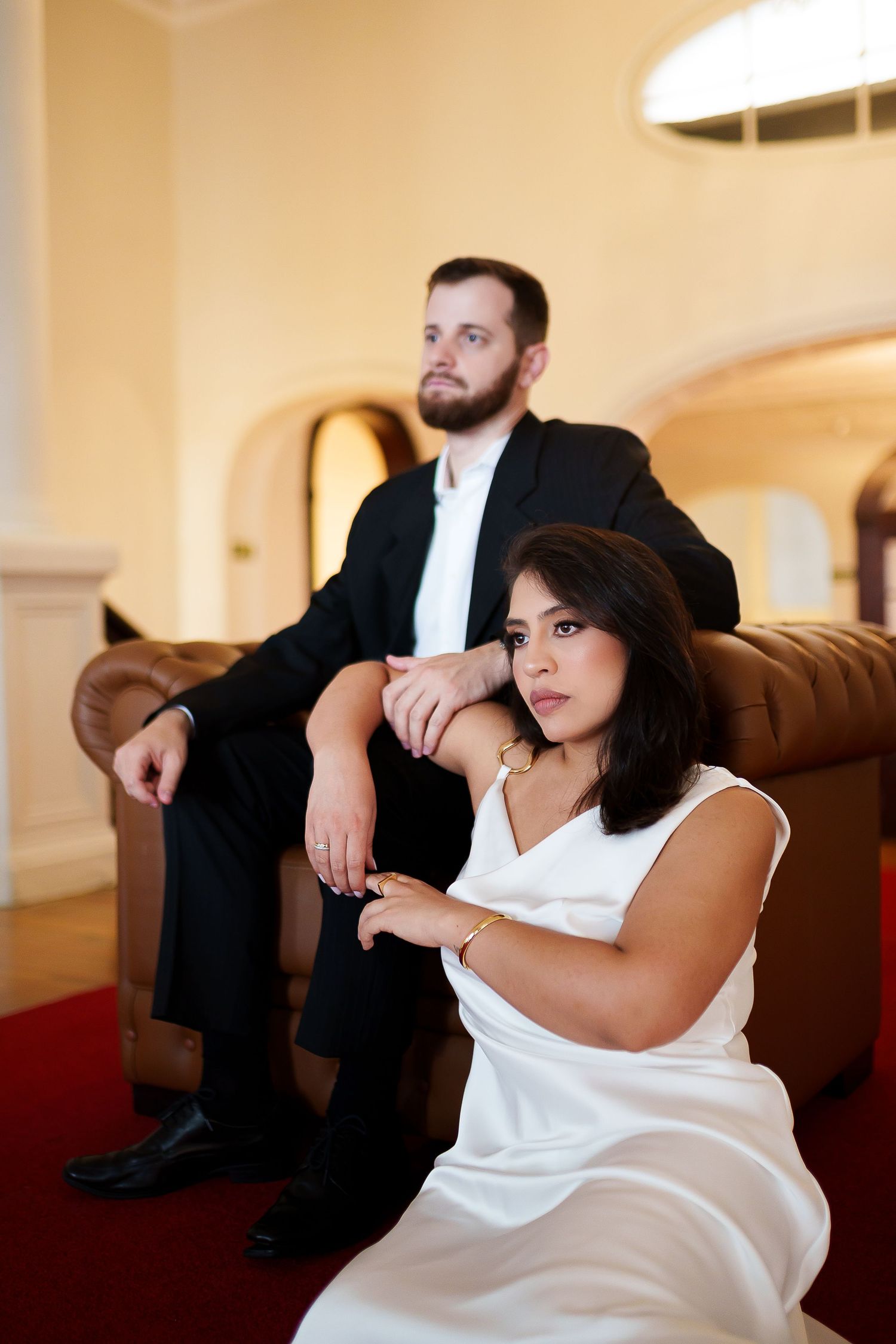 Elegant couple posing in a grand interior, woman in white dress seated on floor, man in black suit on leather sofa.