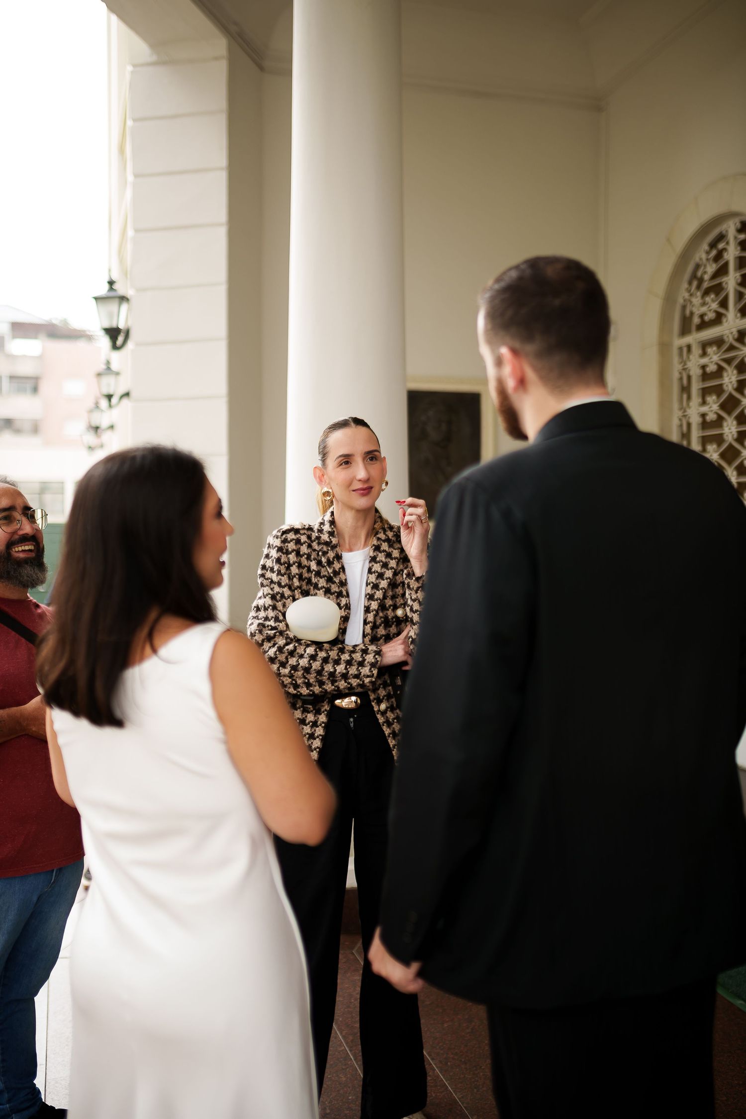 A woman in a houndstooth blazer smiles while conversing with guests at an elegant venue with white columns.
