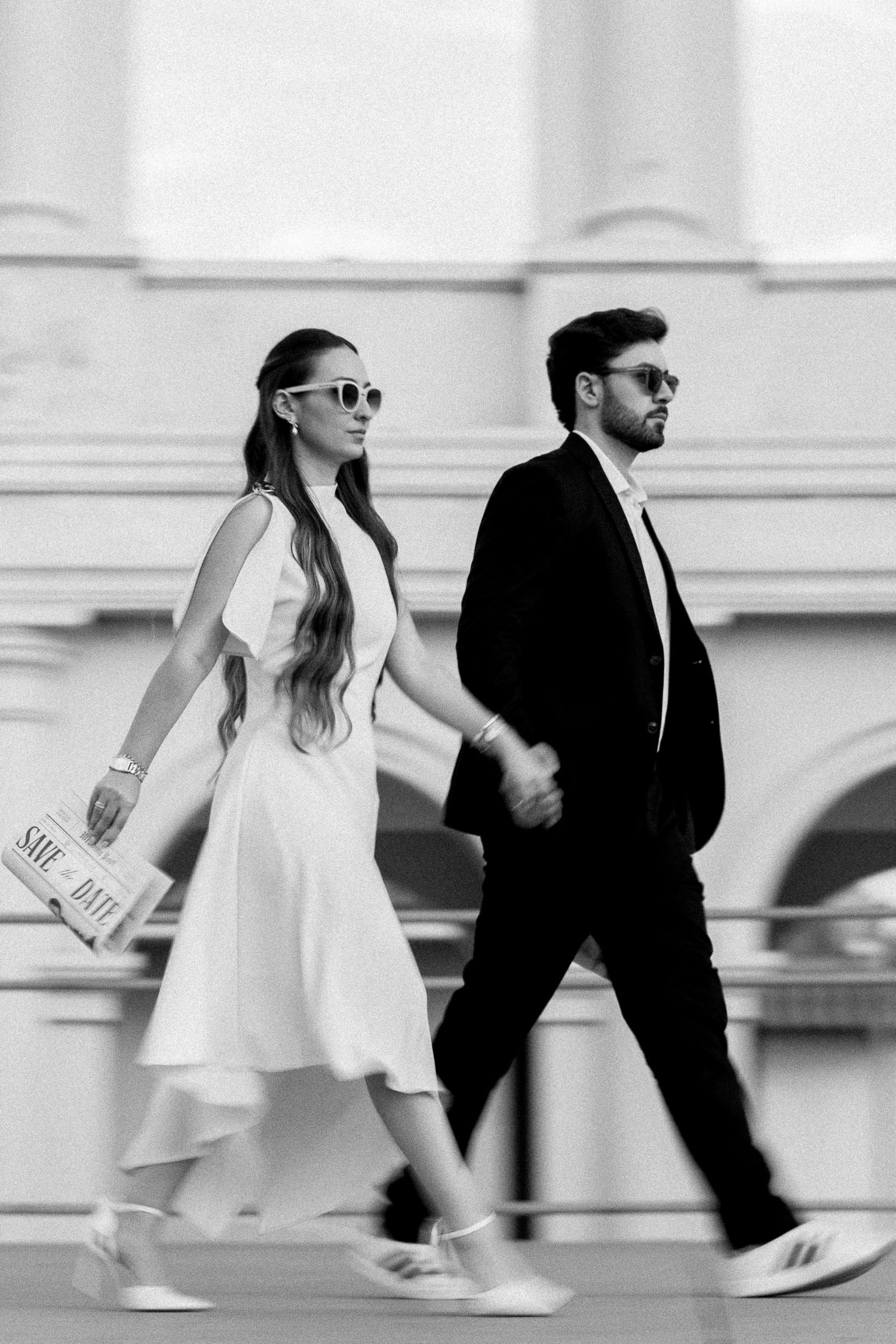 Black and white photo of a stylish couple walking hand in hand, woman in white dress, man in black suit, both wearing sunglasses.