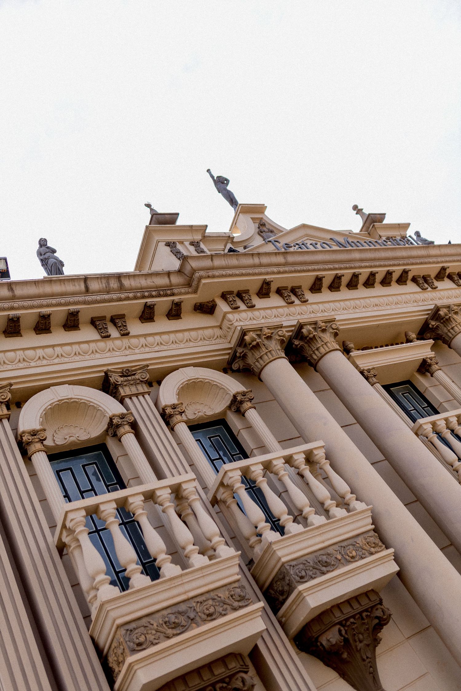 Low-angle view of a neoclassical building facade with ornate columns, balustrades, and statues against a gray sky.