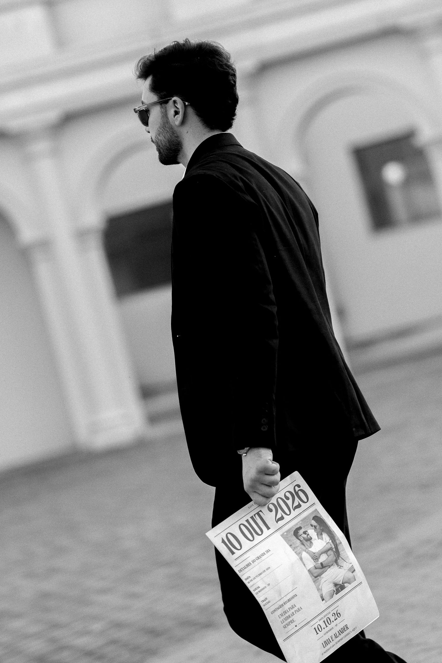 Black and white photo of a man in a suit holding a newspaper, walking outdoors near a classical building.