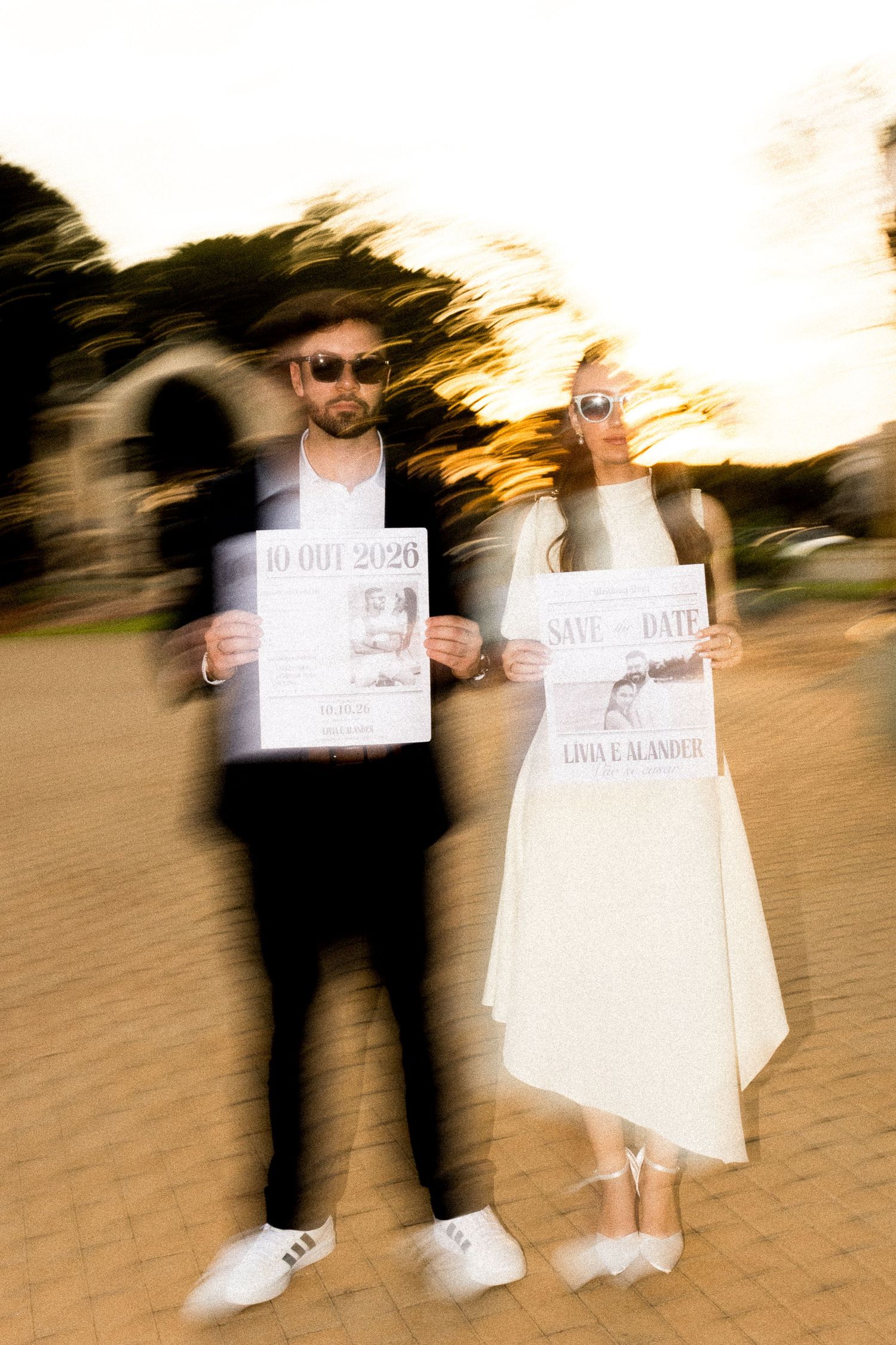 Couple in sunglasses holding Save the Date wedding signs outdoors at dusk with motion blur effect.