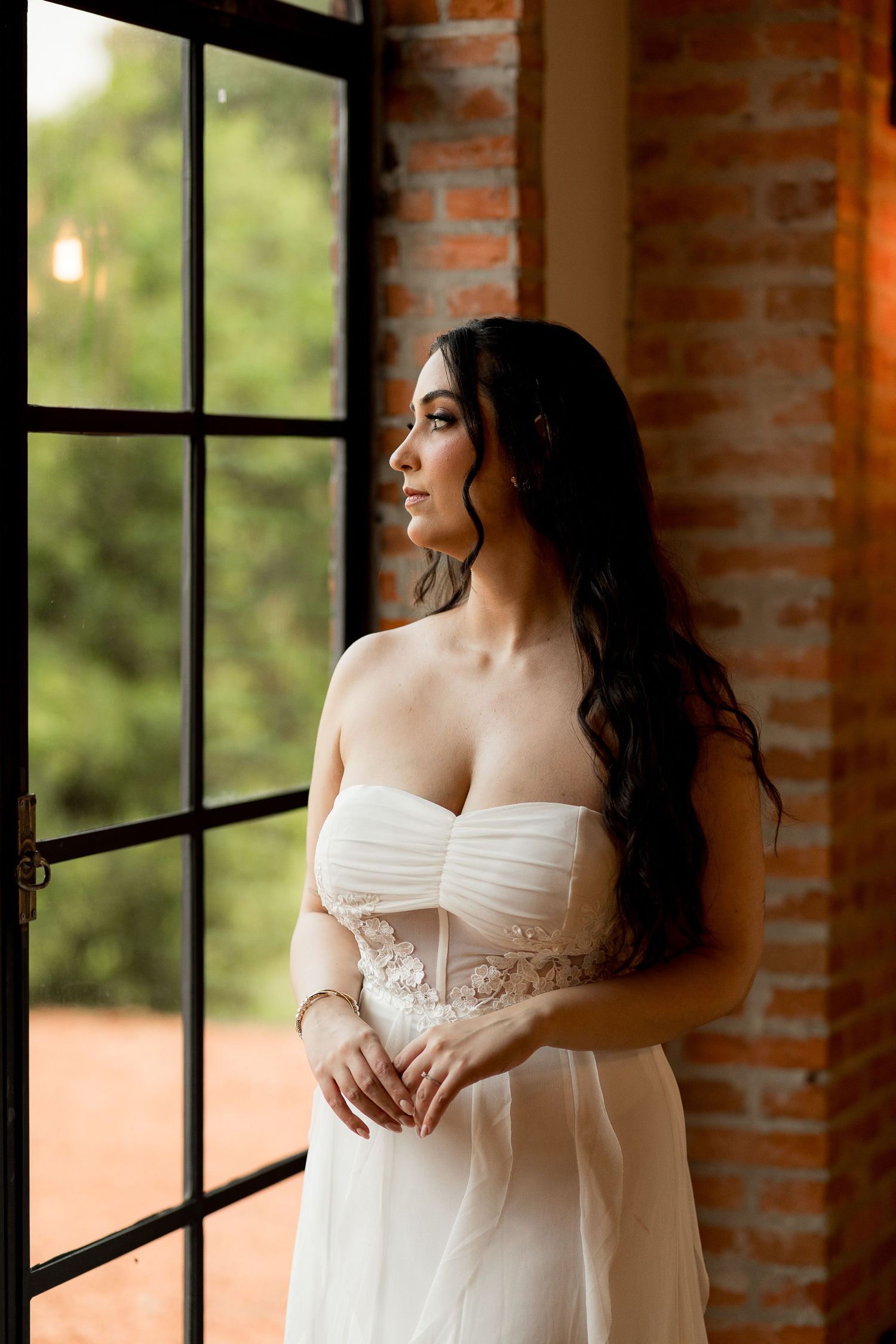 Elegant bride in strapless white lace wedding gown standing by large window with brick wall backdrop.