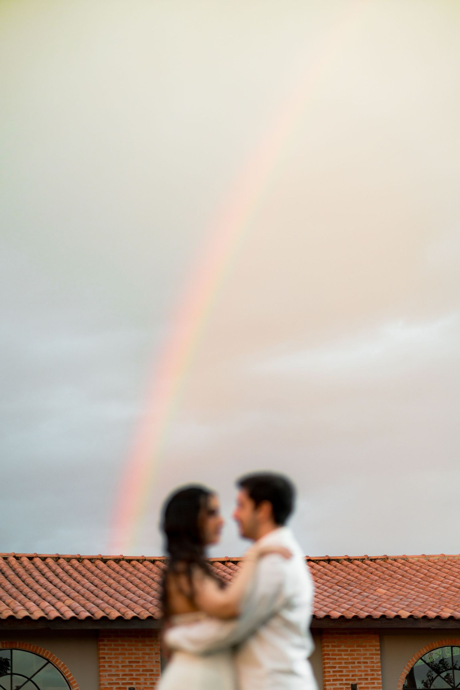 Romantic couple embracing under a beautiful rainbow in a cloudy sky above a terracotta roof building.