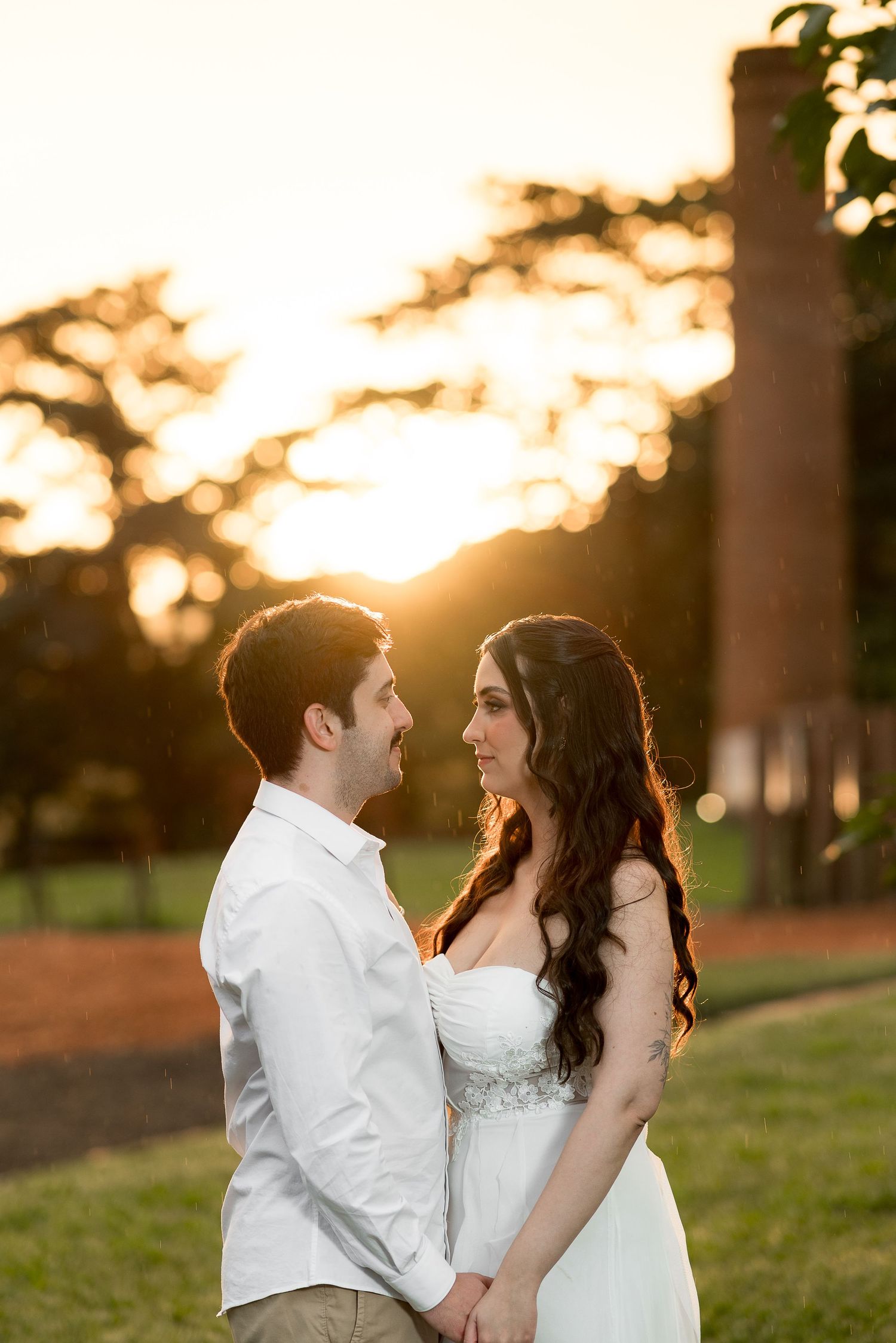 Couple in white attire holding hands at golden hour sunset with bokeh trees and brick chimney backdrop.