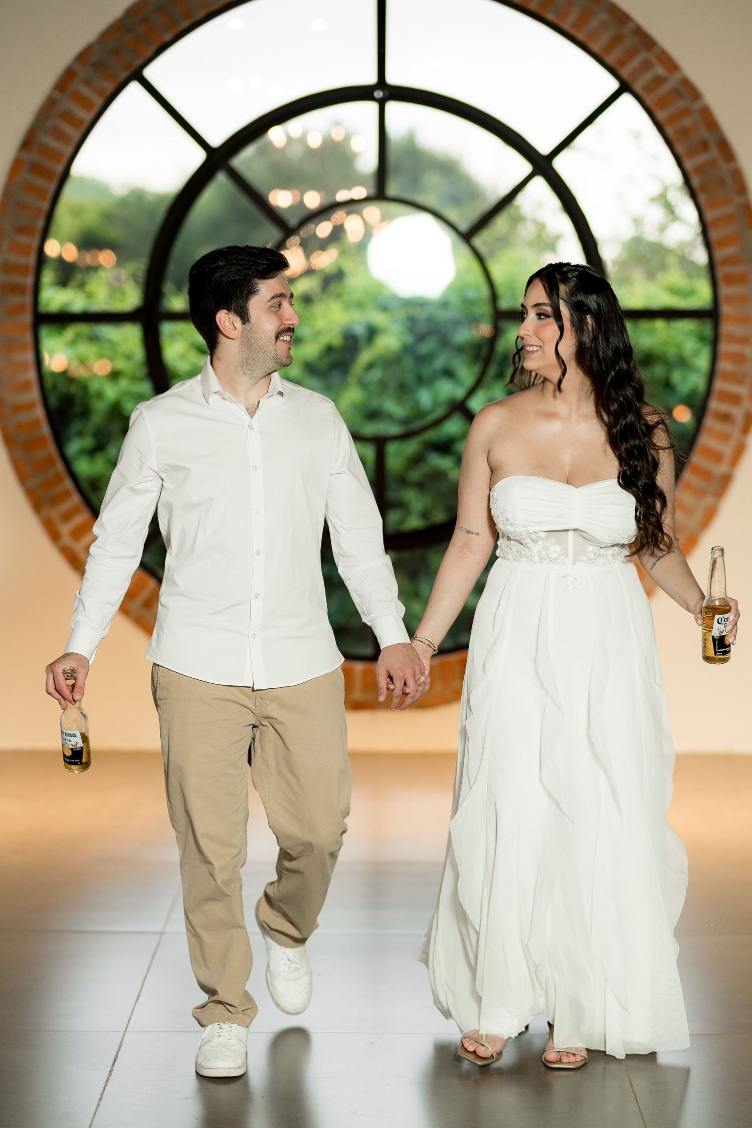 Couple in white outfits holding hands and beer bottles walking in front of a large circular window.