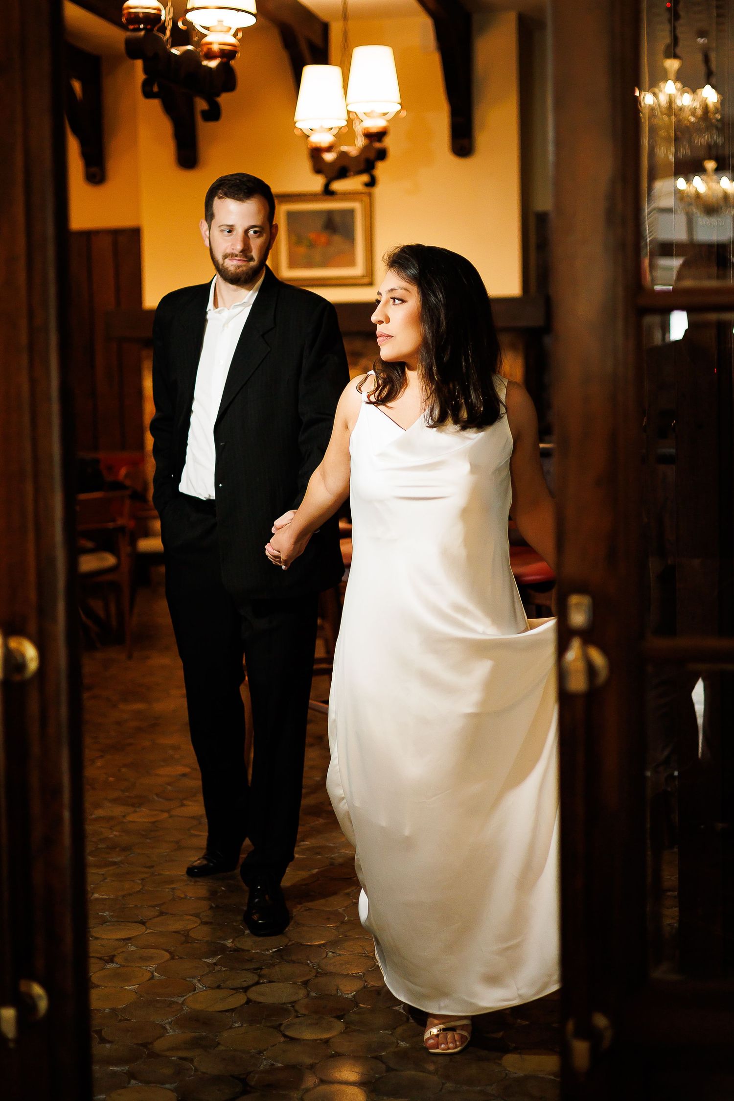 Elegant couple in formal attire holding hands walking through a warmly lit restaurant interior.