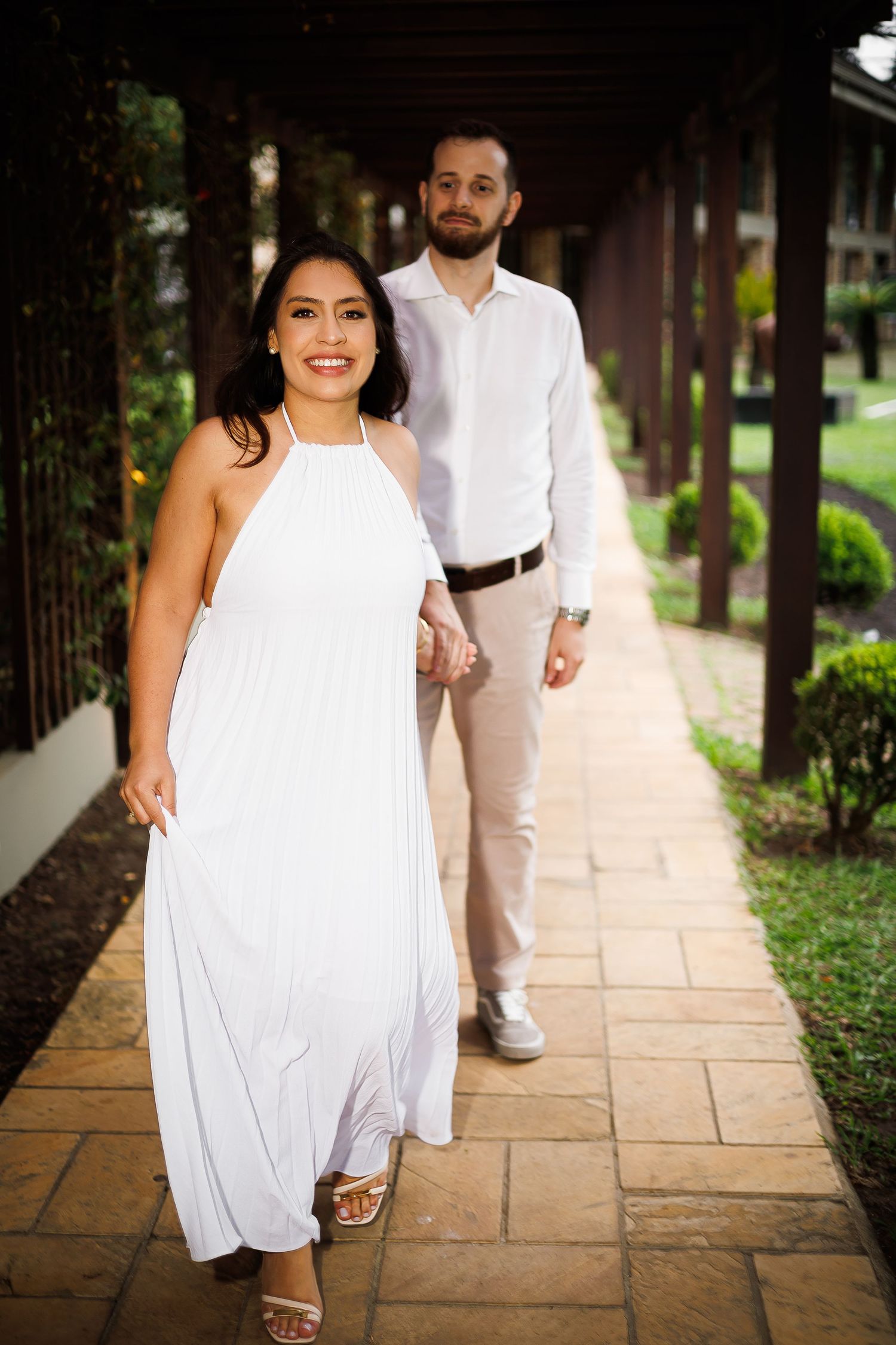 Couple in white outfits walking hand-in-hand along a covered garden pathway, woman in halter dress smiling.