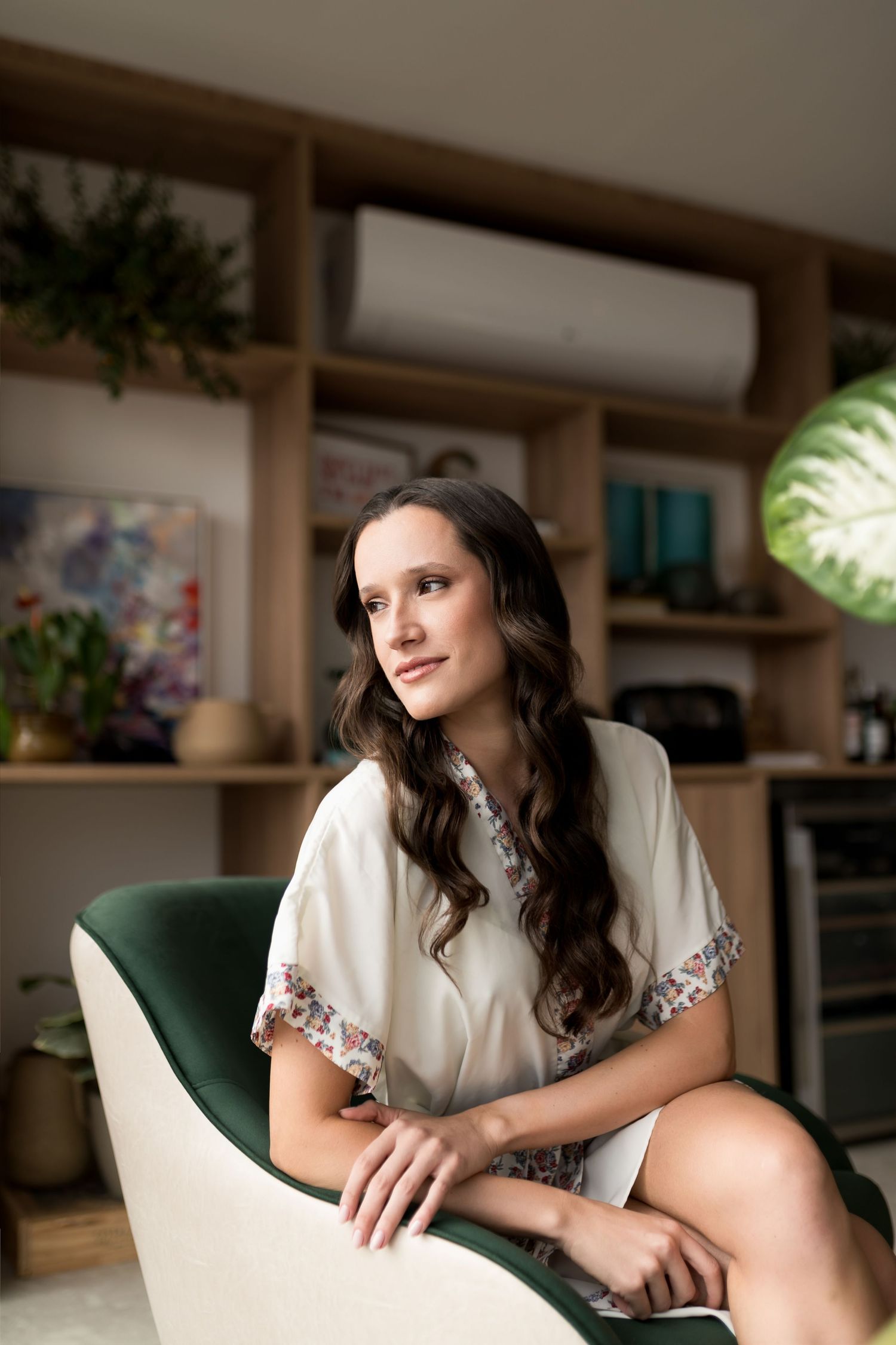 Young woman with long dark hair sits in a green velvet chair, wearing a floral dress in a modern home interior.