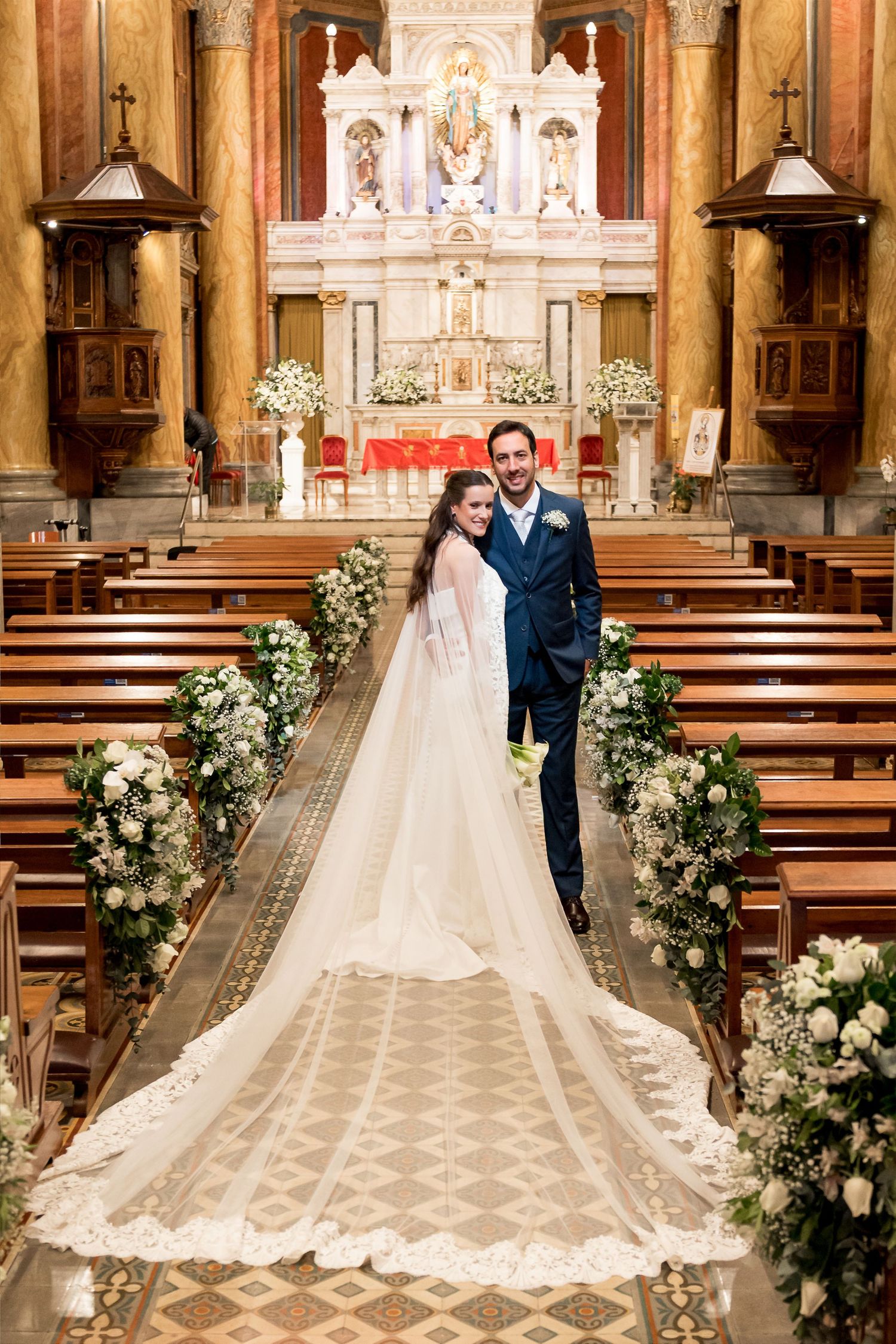 Newlyweds stand together in ornate church aisle lined with white floral arrangements, bride in elegant gown with cathedral veil.