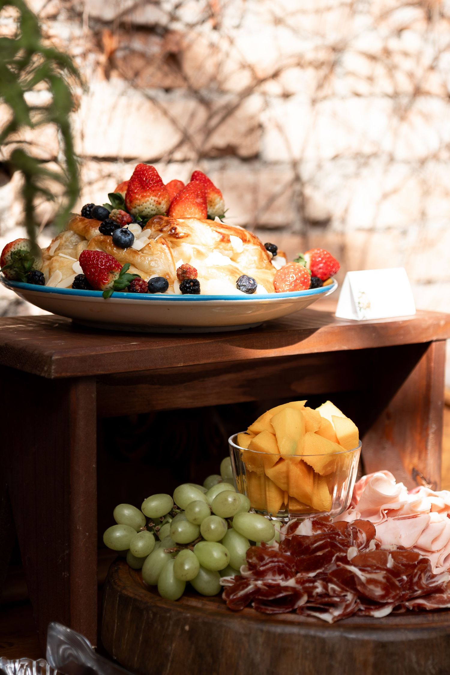 Elegant charcuterie display with brie topped with fresh berries, green grapes, cantaloupe, and cured meats on wooden shelves.