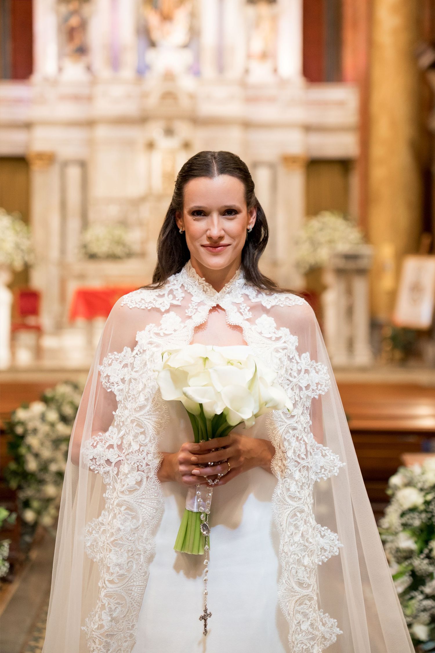 Bride in white lace cape gown holding white rose bouquet stands before church altar on wedding day.