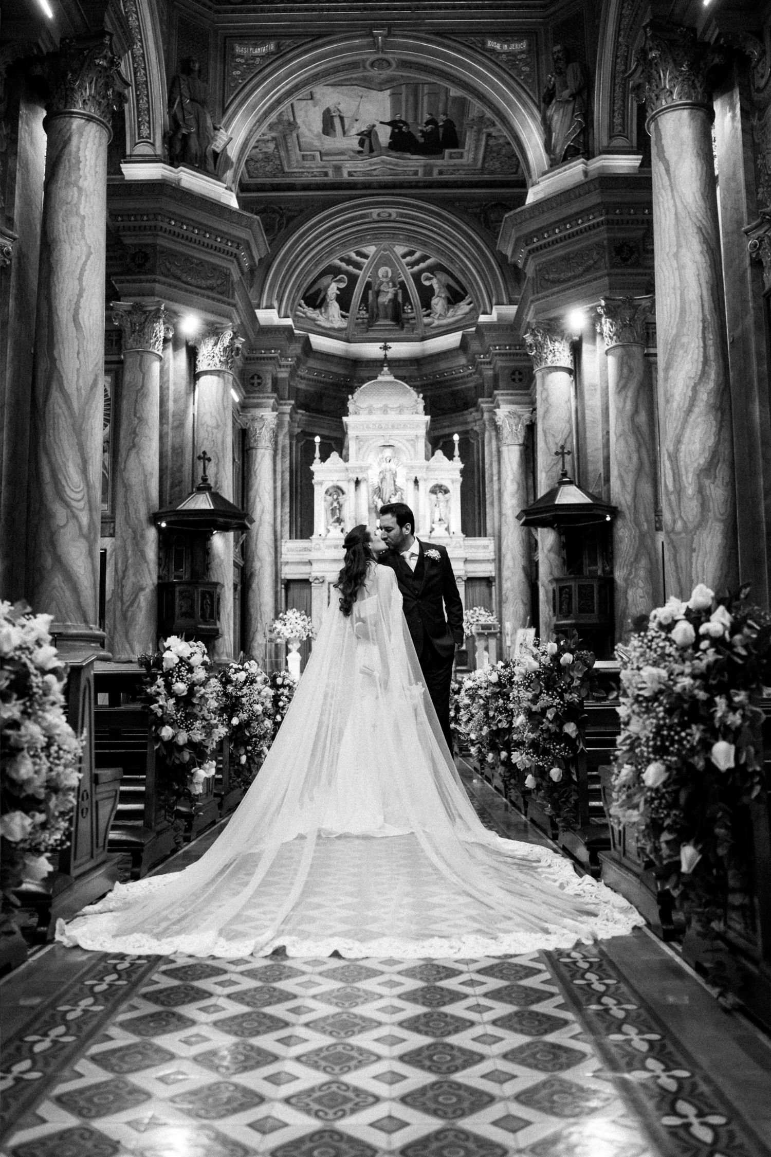 Black and white photo of bride and groom kissing at ornate church altar, bride's long veil trailing down the aisle.