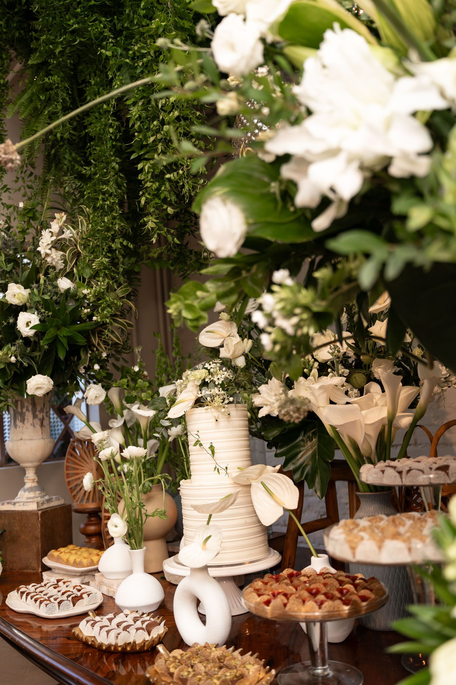 Elegant wedding dessert table with white tiered cake, floral arrangements, and assorted sweets surrounded by lush greenery.