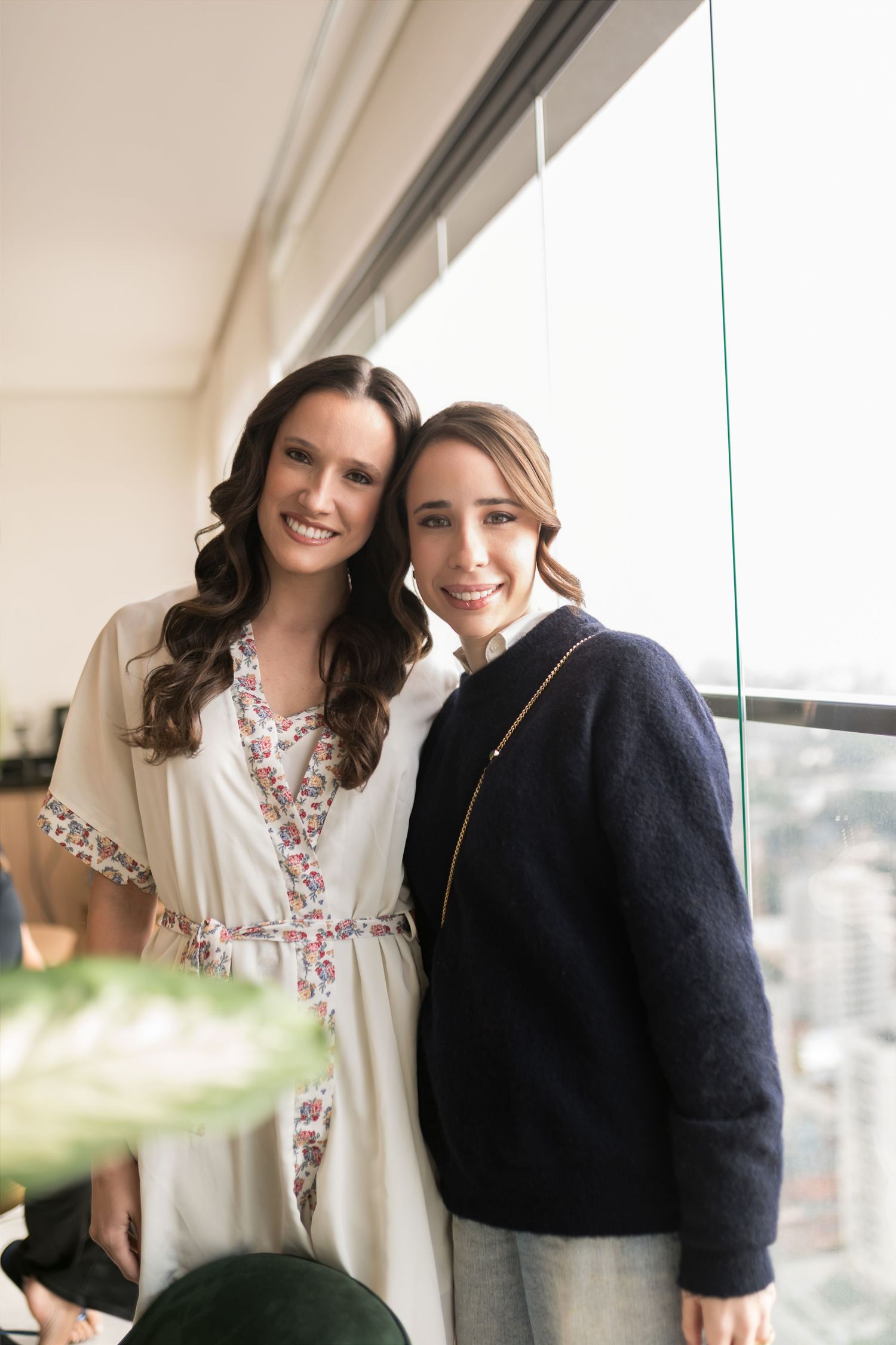 Two smiling women posing together near a large window, one in a floral robe and one in a navy sweater.