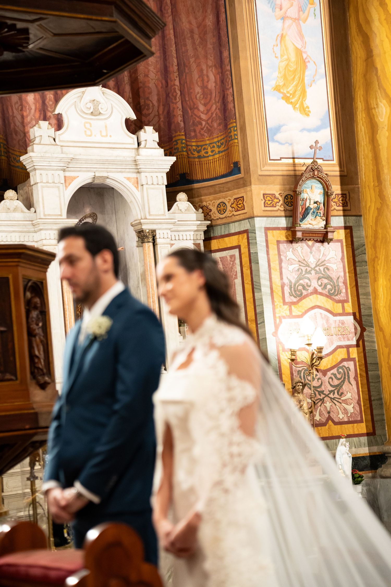 Bride in lace gown and groom in blue suit stand at the altar of an ornate church during wedding ceremony.